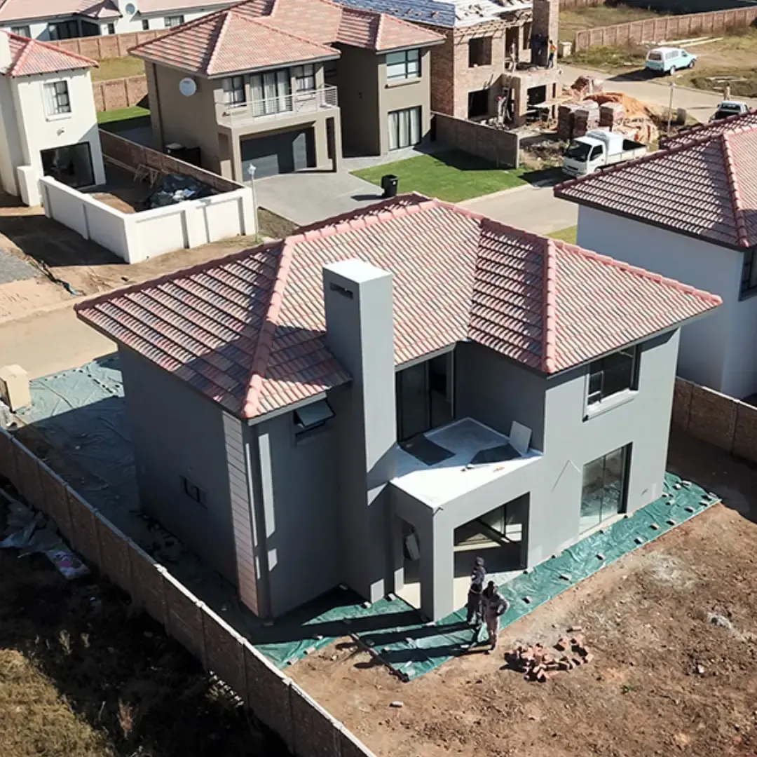 Gray two-story house with red roof, chimney, and construction materials. Two people stand outside.