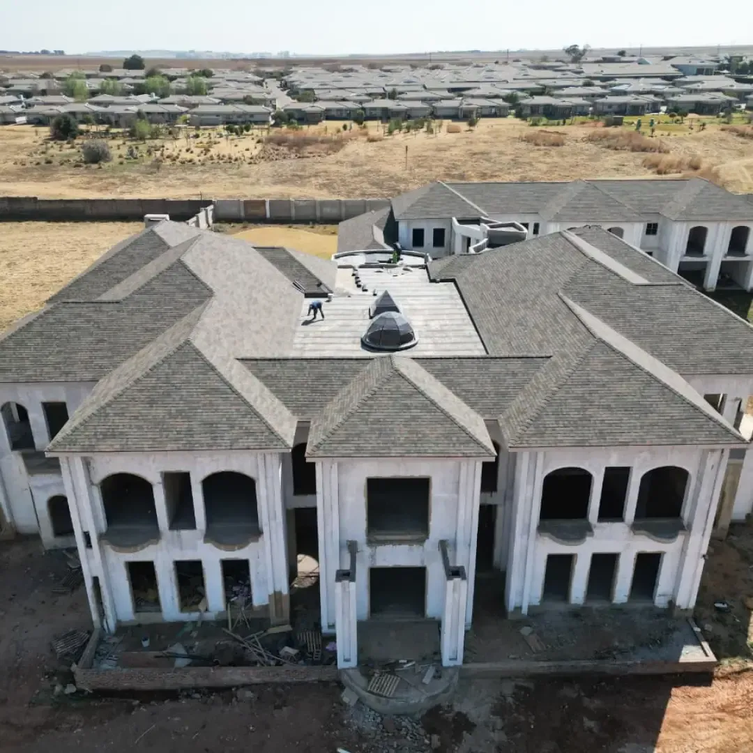 Construction of a large two-story house with gray roof, set in a rural area with other houses in the background.