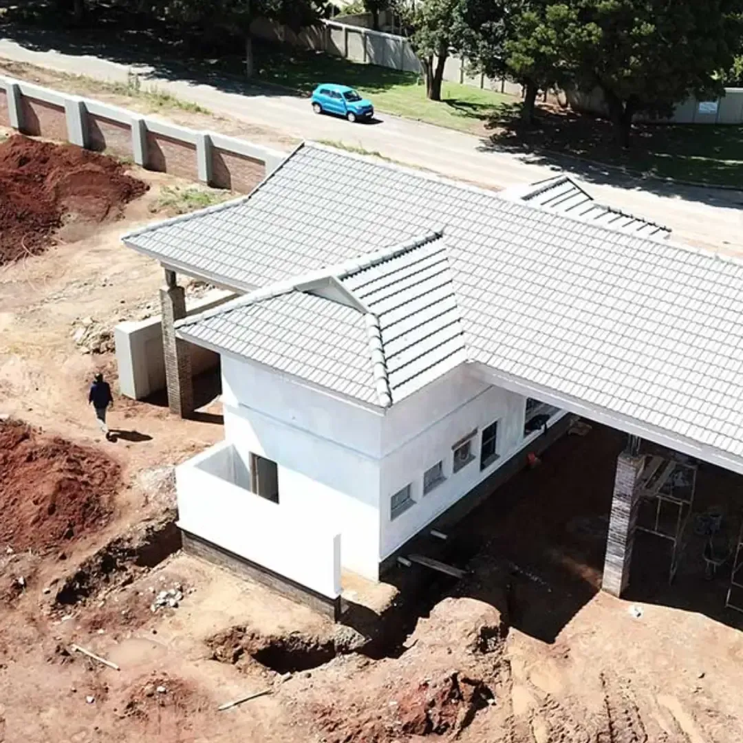 Aerial view of a white building with gray tiled roof under construction. A person walks nearby, a blue car passes on the road.