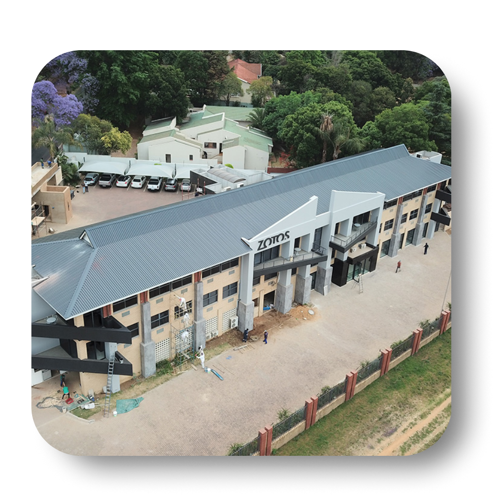 Aerial view of a modern building with gray roof, beige walls, and dark windows; surrounded by trees and a fence.