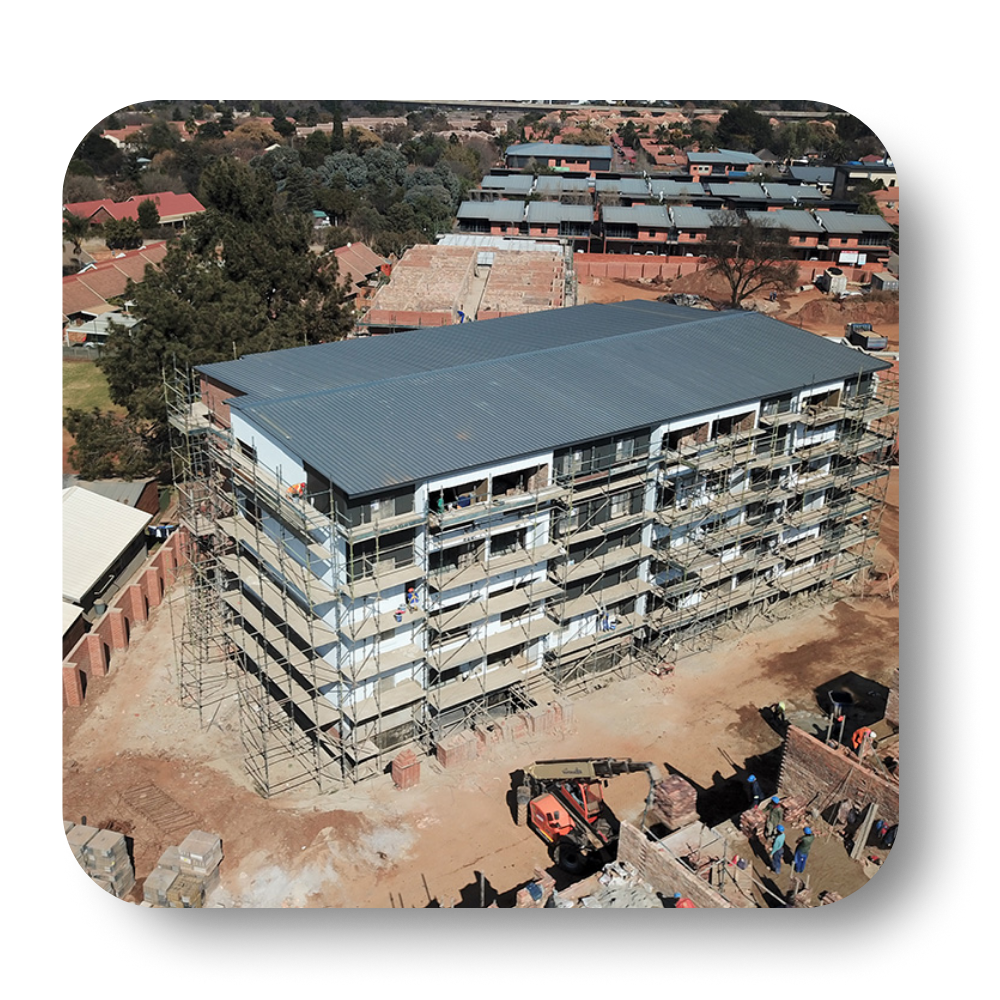 Aerial view of a multi-story building under construction; scaffolding surrounds the facade, roof is complete, dusty environment.