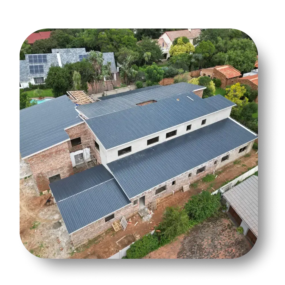 Aerial view of a building under construction with a dark gray roof, surrounded by trees and other houses.