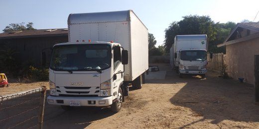 Two white moving trucks parked in a dirt driveway between buildings on a sunny day.