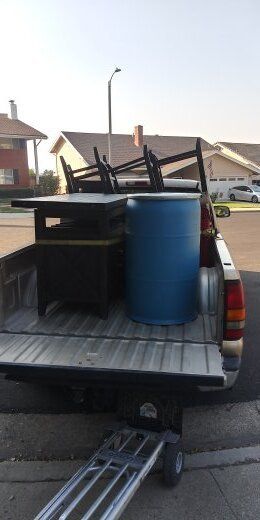 A pickup truck bed is loaded with a black table, a blue barrel, and a hand truck on a suburban street.