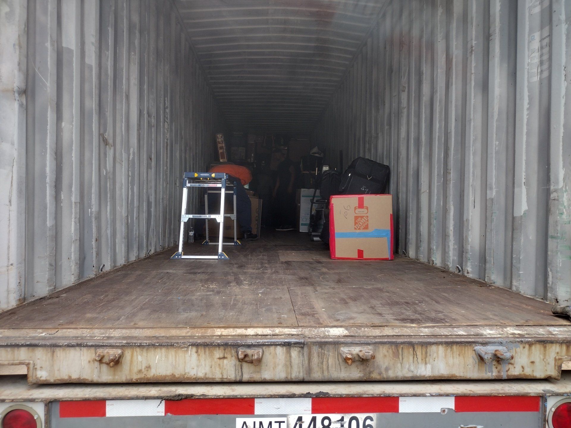 Inside a shipping container, items sit on a wooden floor. A person is visible near the back, among miscellaneous objects.