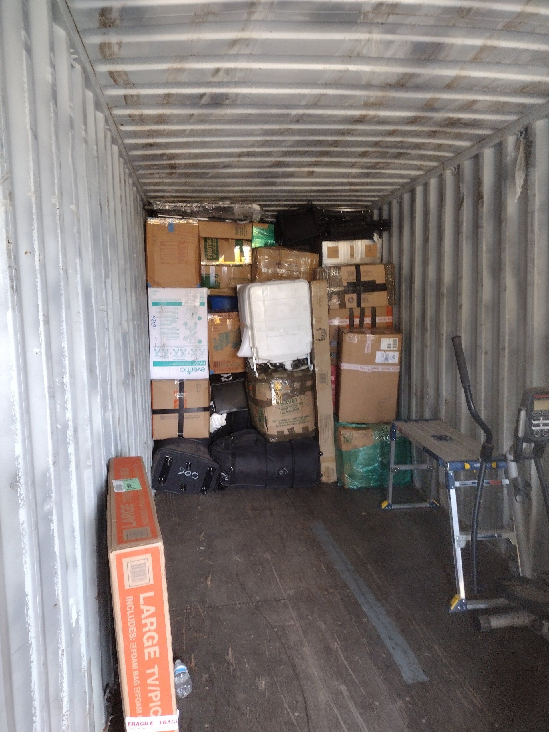Inside a cluttered metal shipping container. Boxes, furniture, and equipment are densely packed, viewed from the entrance.