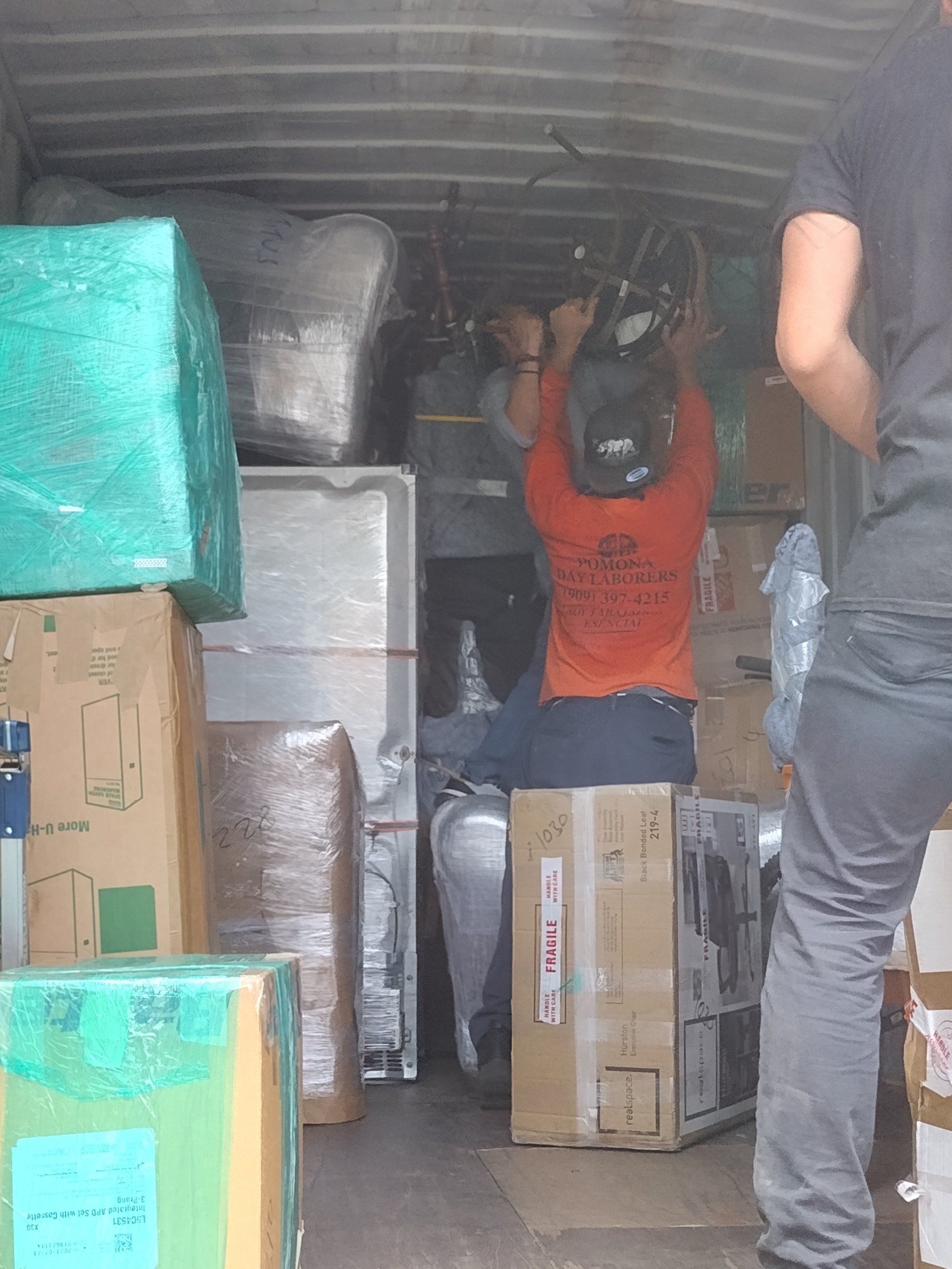 Inside a truck being loaded. A person in orange shirt adjusts object above them. Many packed boxes are present.