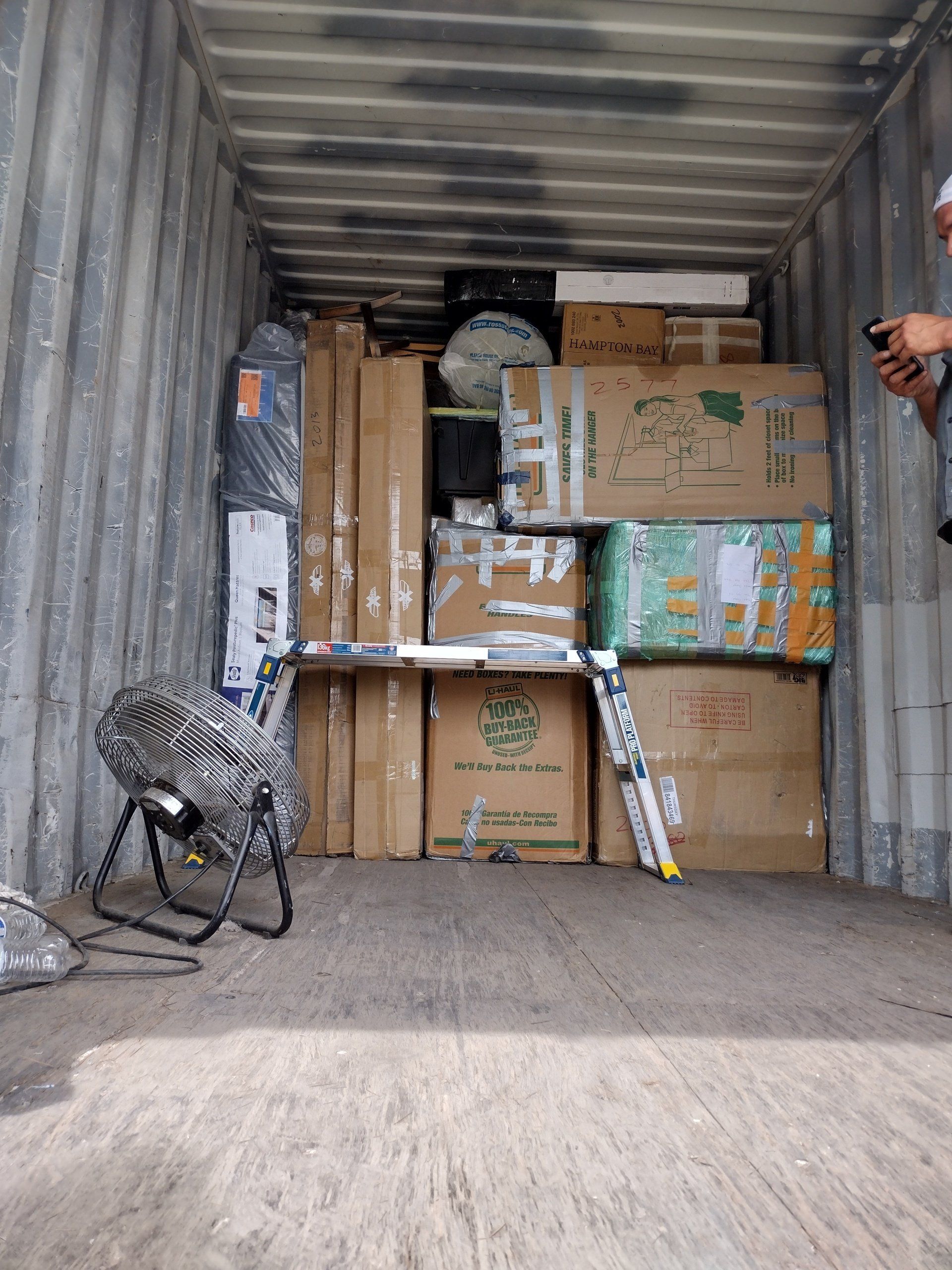 Inside shipping container filled with packed cardboard boxes, fan, ladder, and various items. Man takes photo.