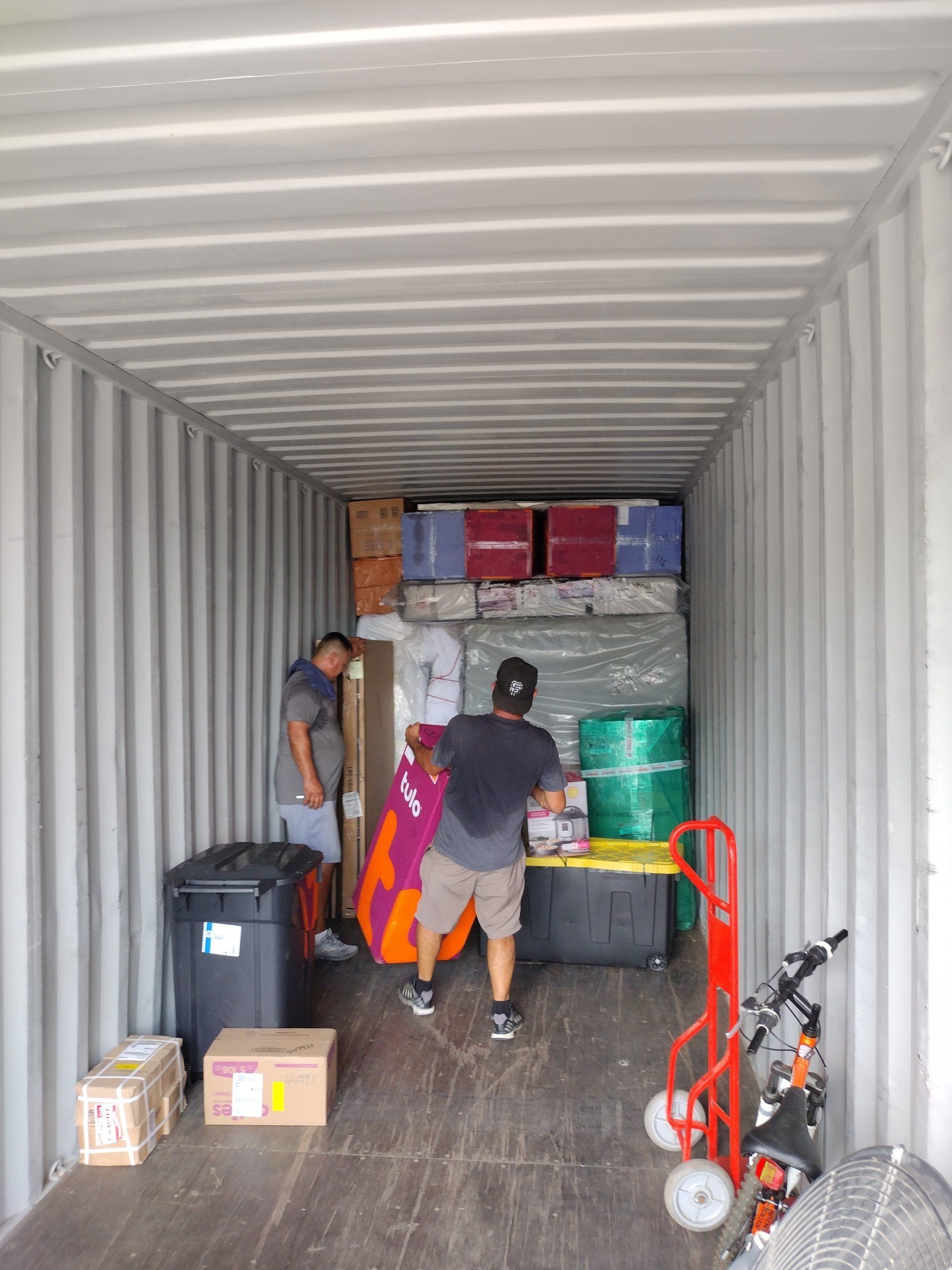 Two men loading a storage container with boxes, furniture, and supplies. Inside a shipping container.