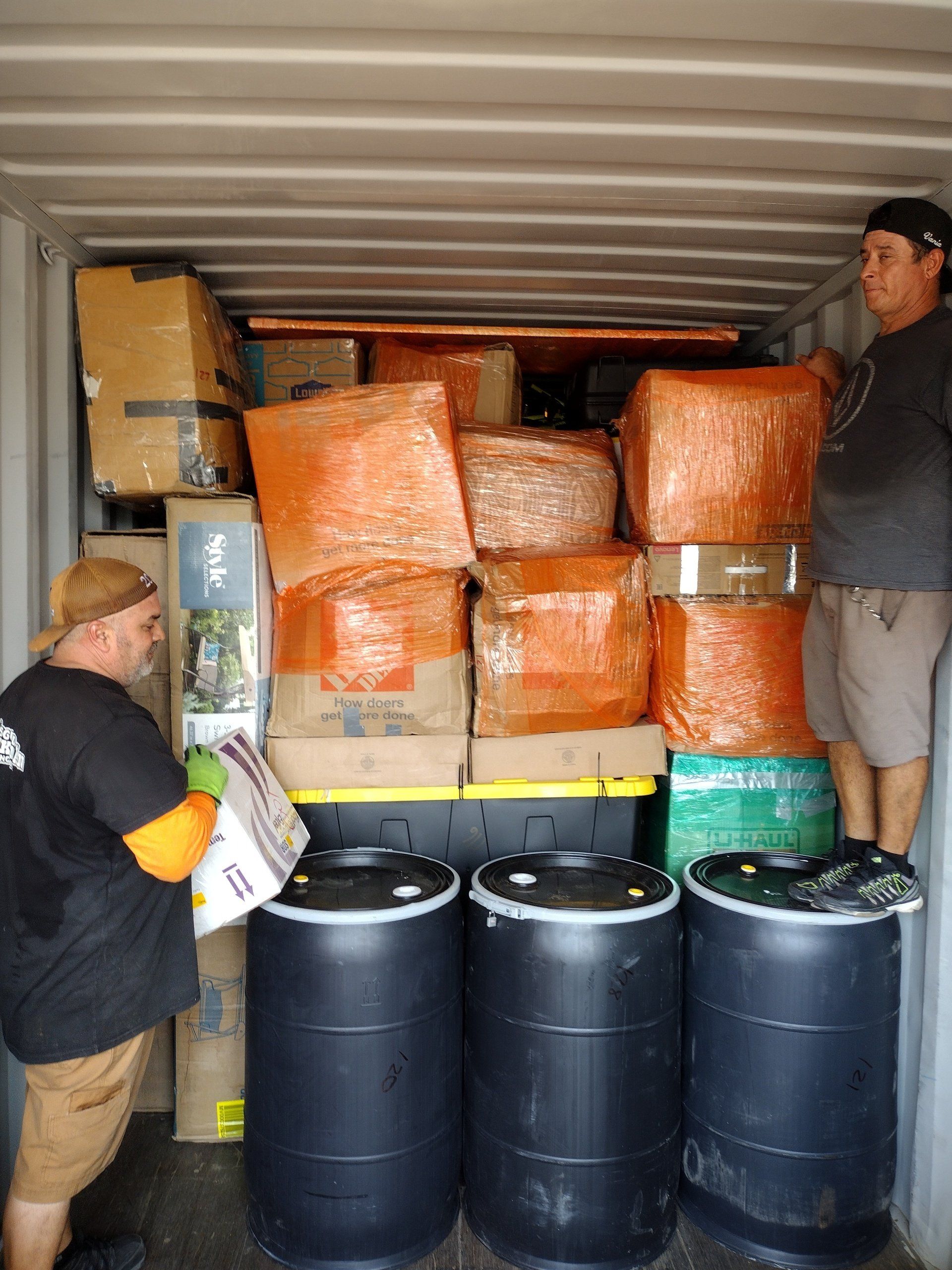 Two men packing boxes and barrels inside a shipping container.
