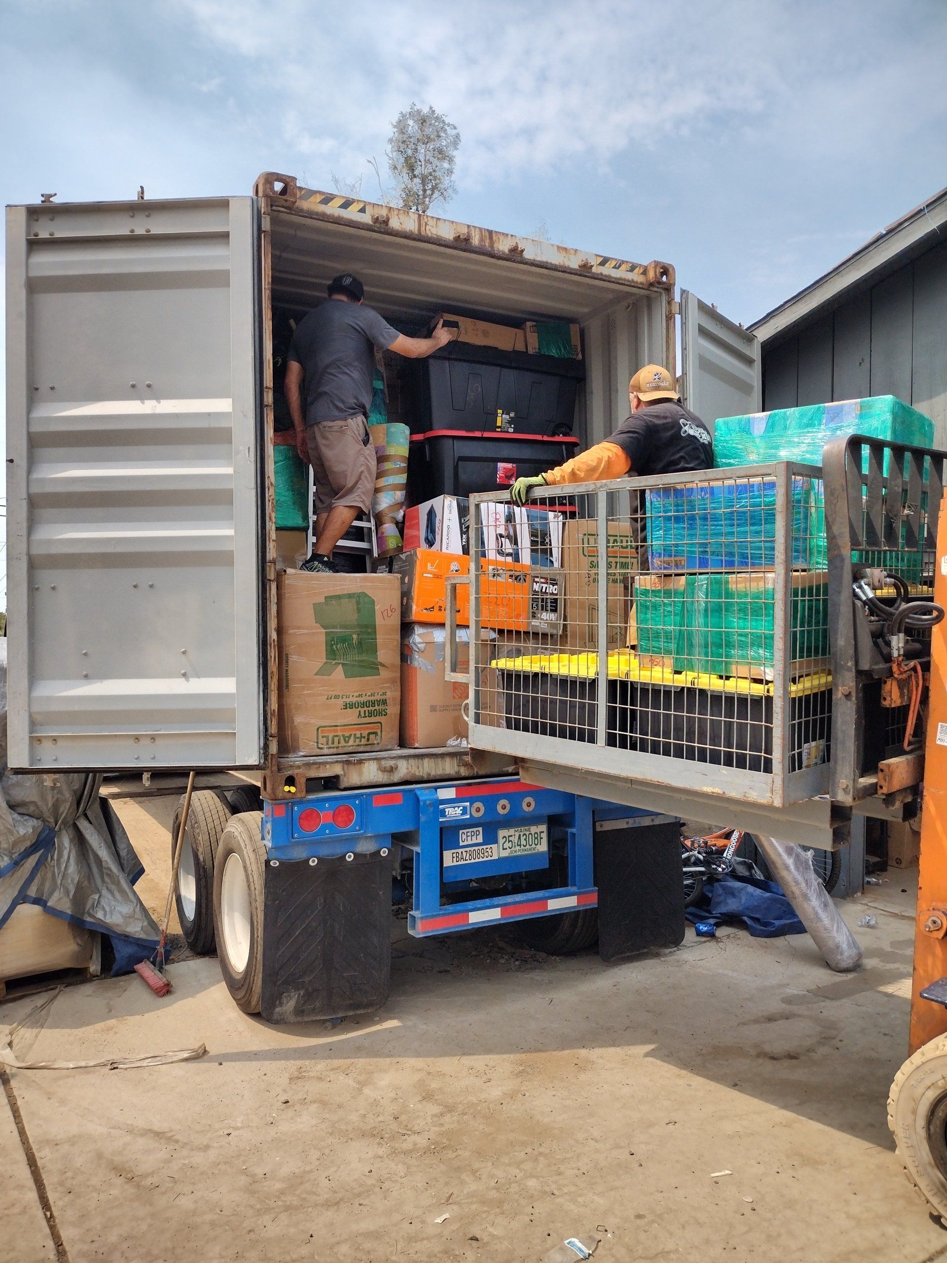 Men loading supplies into a shipping container on a truck, outdoors. Boxes, water bottles, and equipment are being organized.
