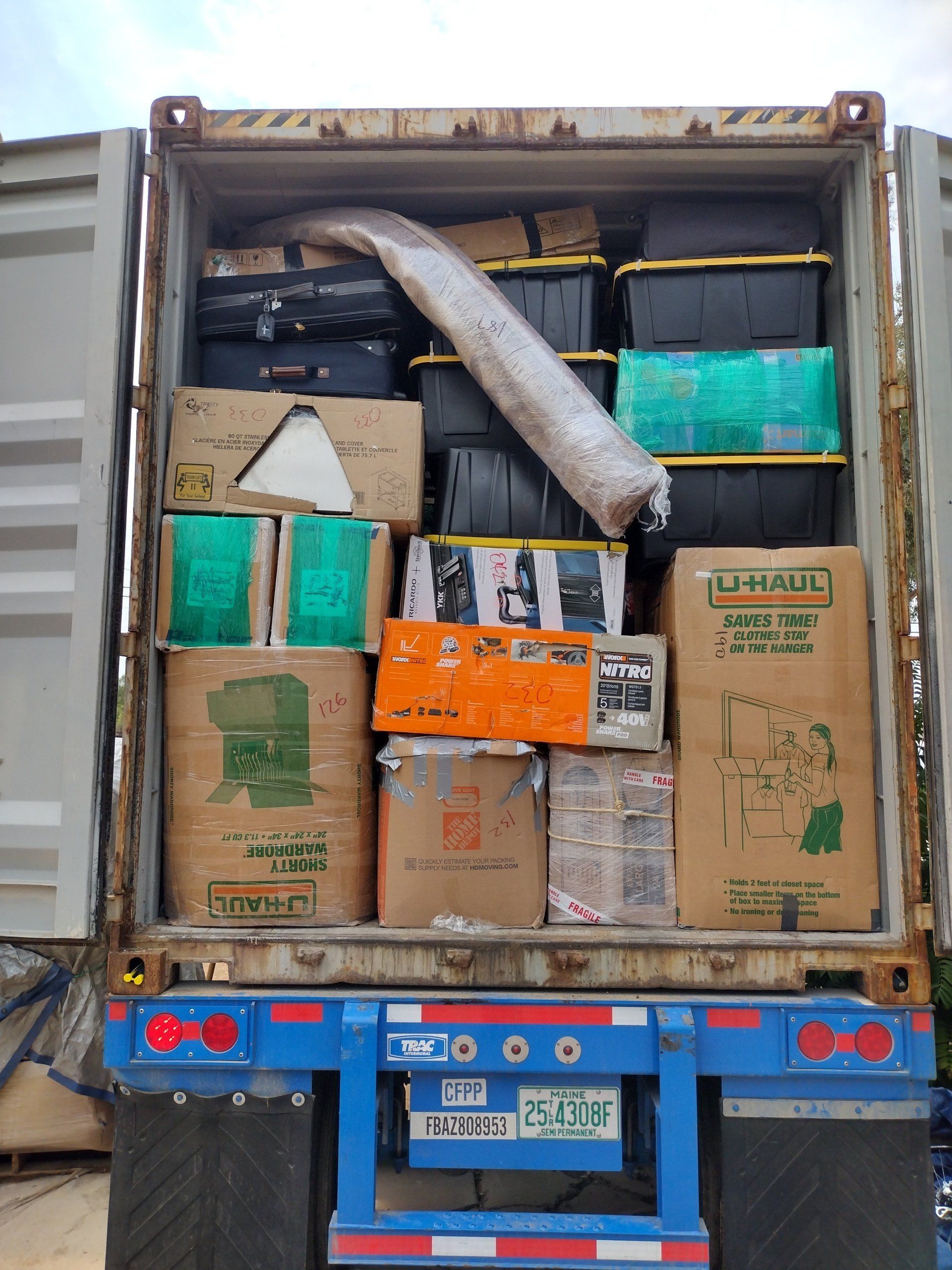 Shipping container filled with boxes, containers, and wrapped items, viewed from the open door of the container.