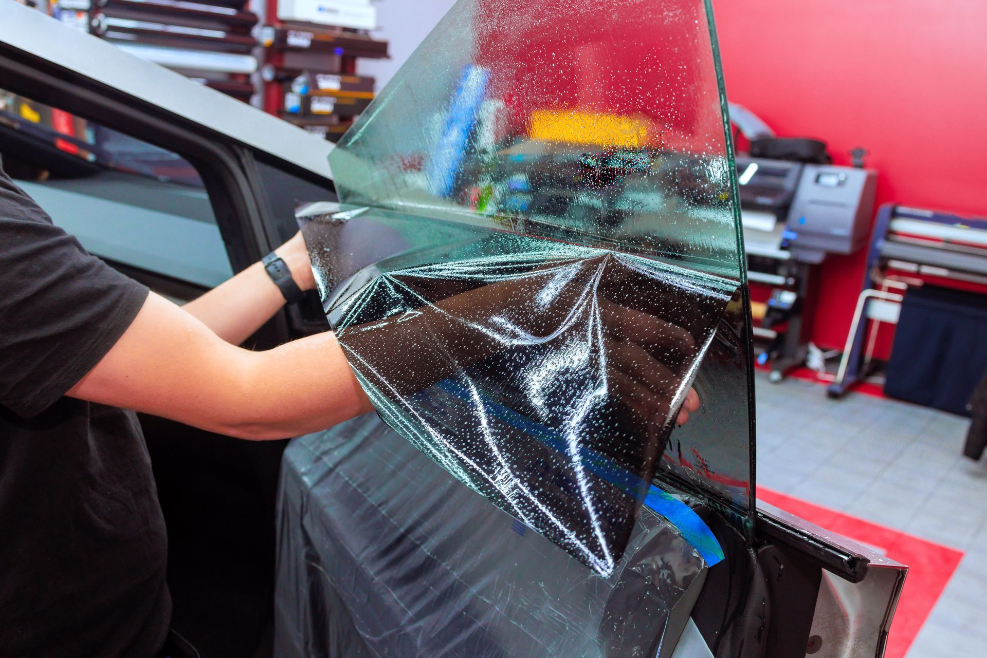 A technician in a shop applies dark, adhesive window tinting film to a vehicle's side window.
