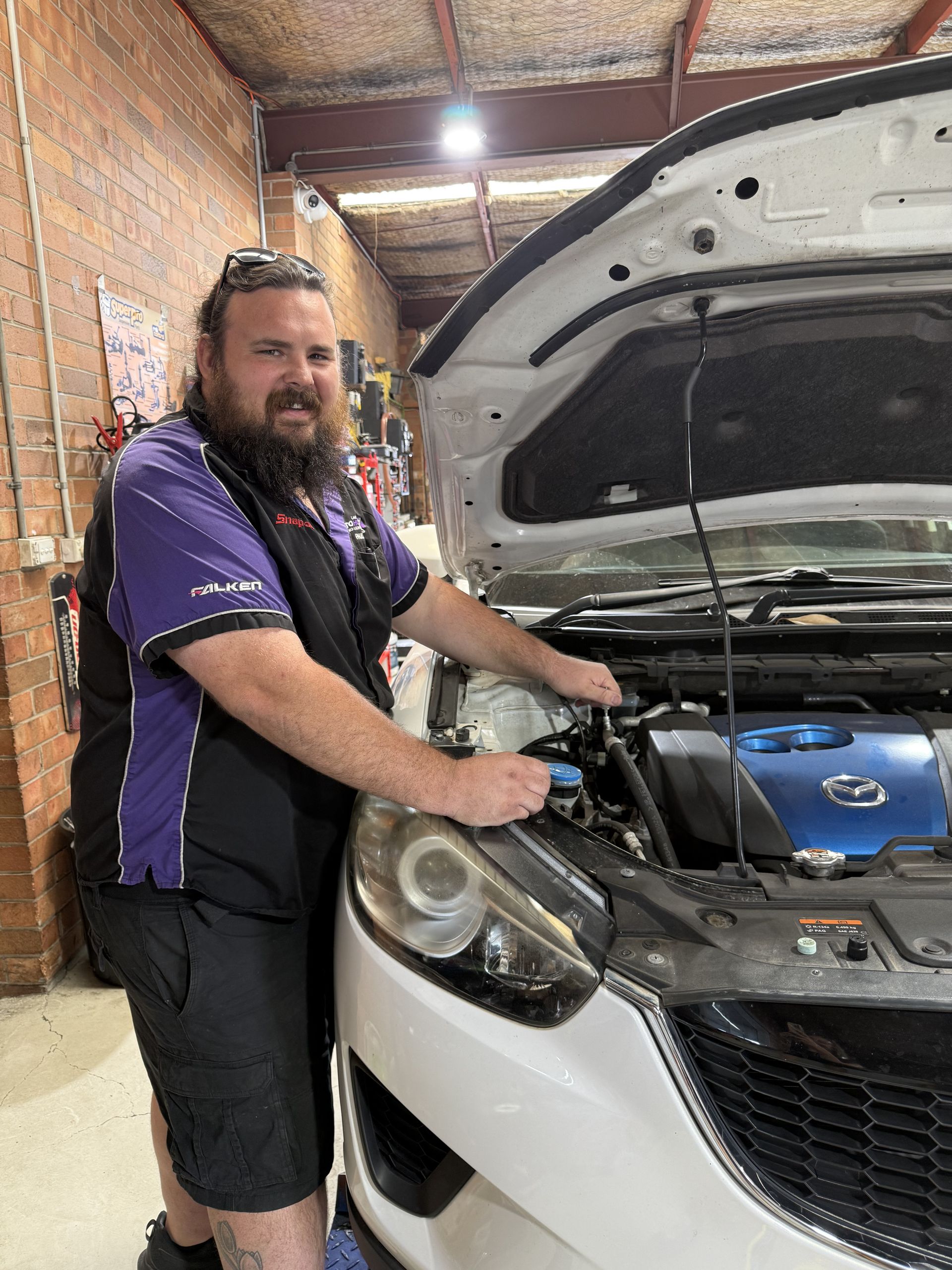 Mechanic in purple and black uniform working on the engine of a white car in a garage— Servicing And Repairs in Lambton, NSW