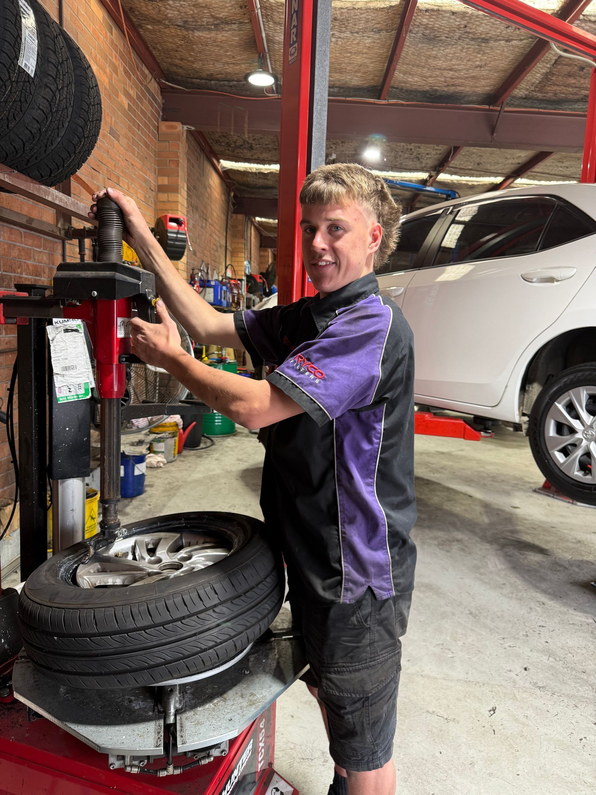 Mechanic working with tire machine in a garage. Wearing black shorts and purple shirt, he smiles— Servicing And Repairs in Lambton, NSW