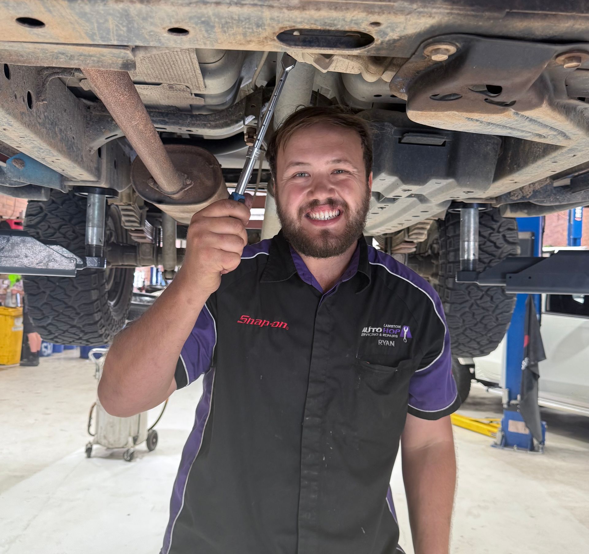 Mechanic smiling under a vehicle, holding a wrench. Wearing a black and purple uniform. Shop setting— Servicing And Repairs in Lambton, NSW