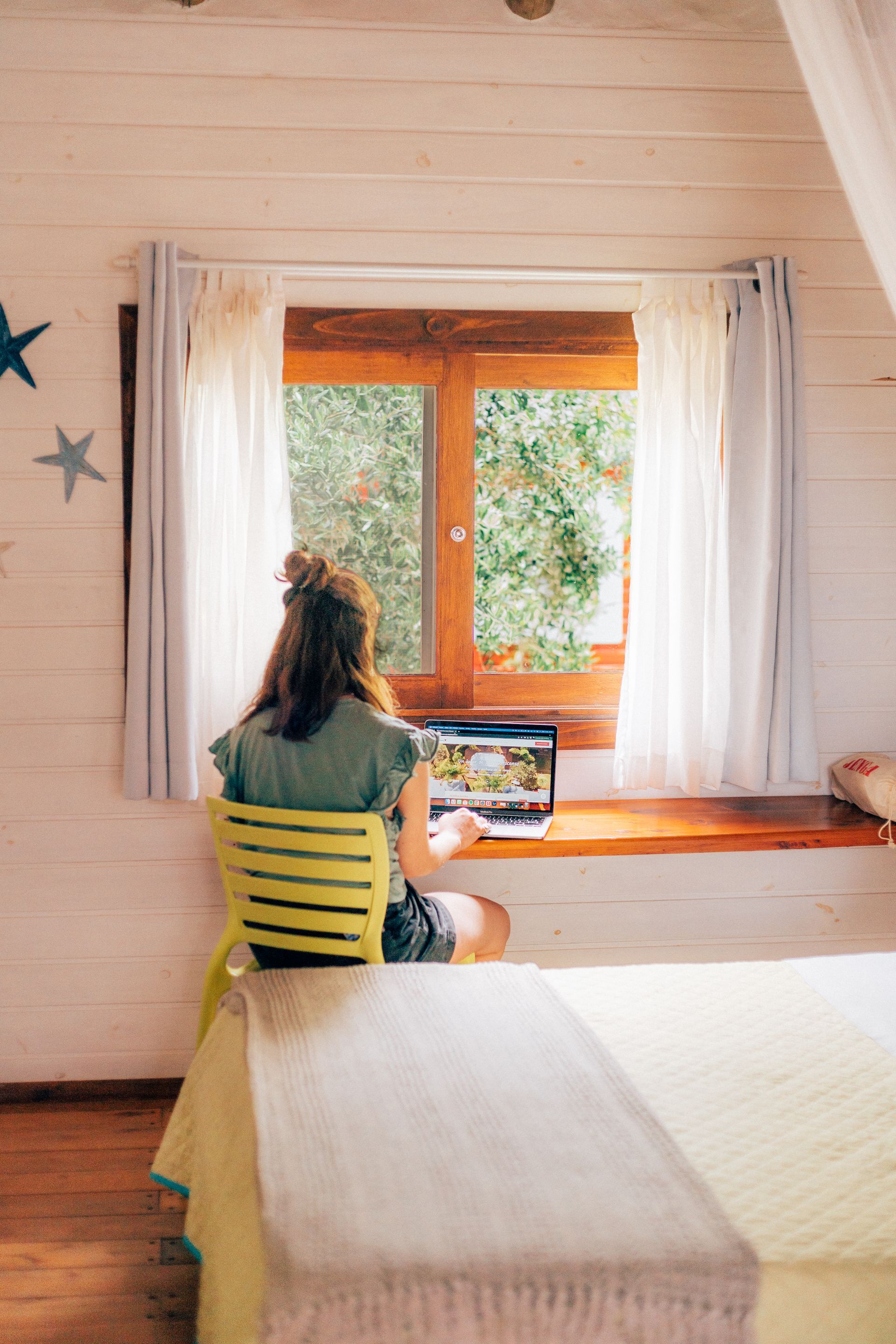 A woman is sitting at a desk in front of a window using a laptop computer.