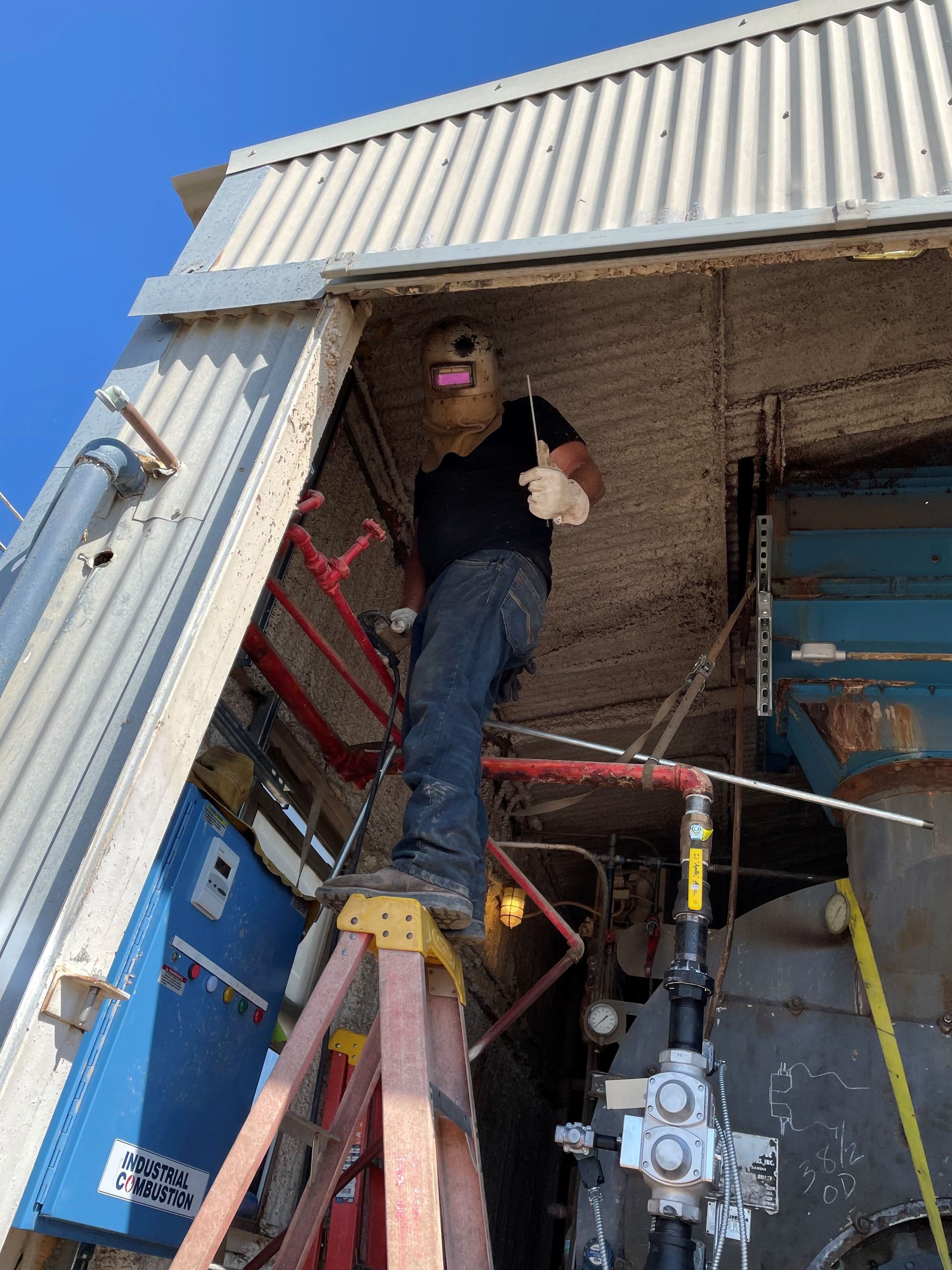 Person in a mask giving thumbs up, standing on a ladder inside a industrial building.