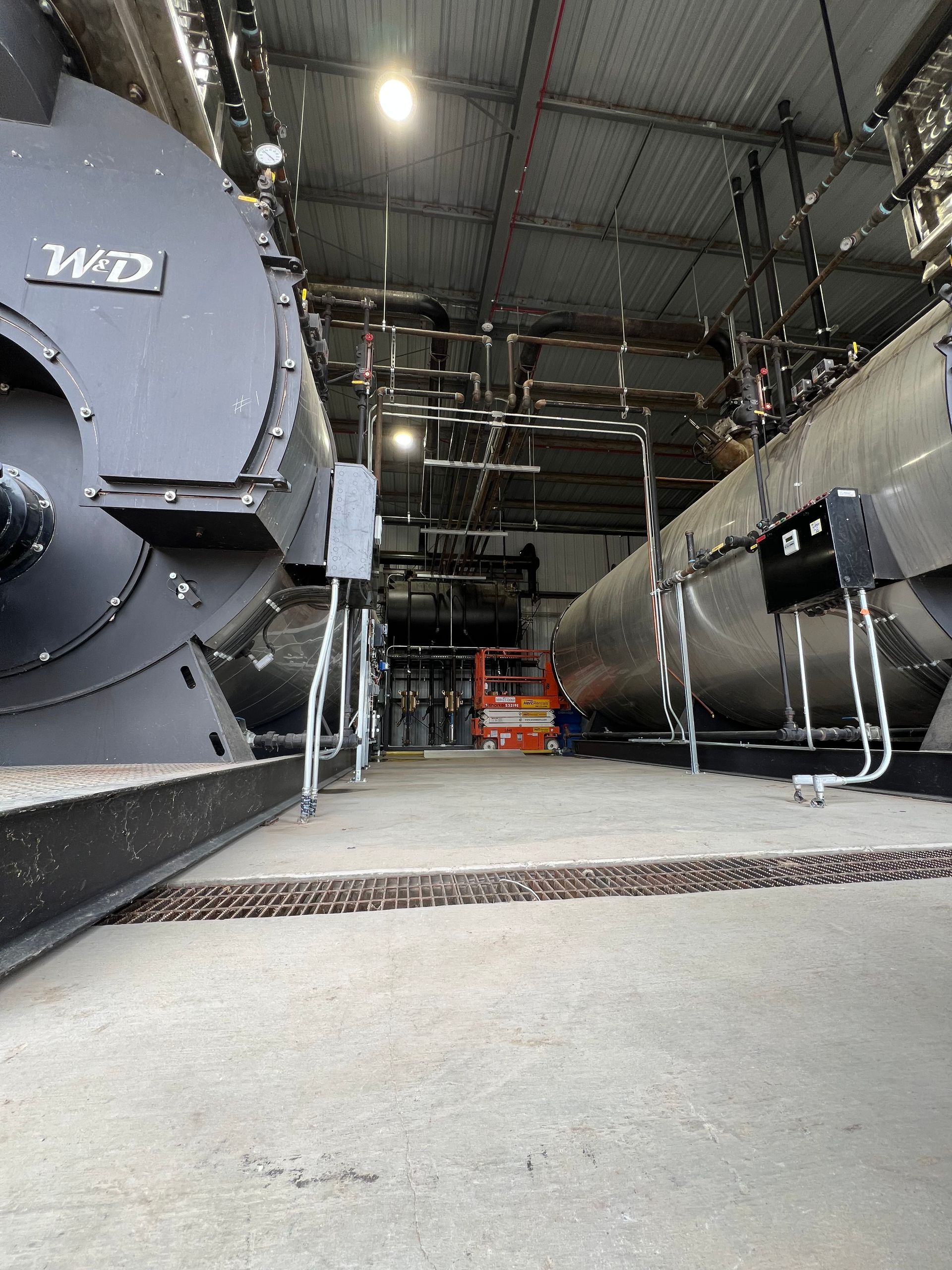 Two large industrial boilers in a factory setting, with pipes and a concrete floor.