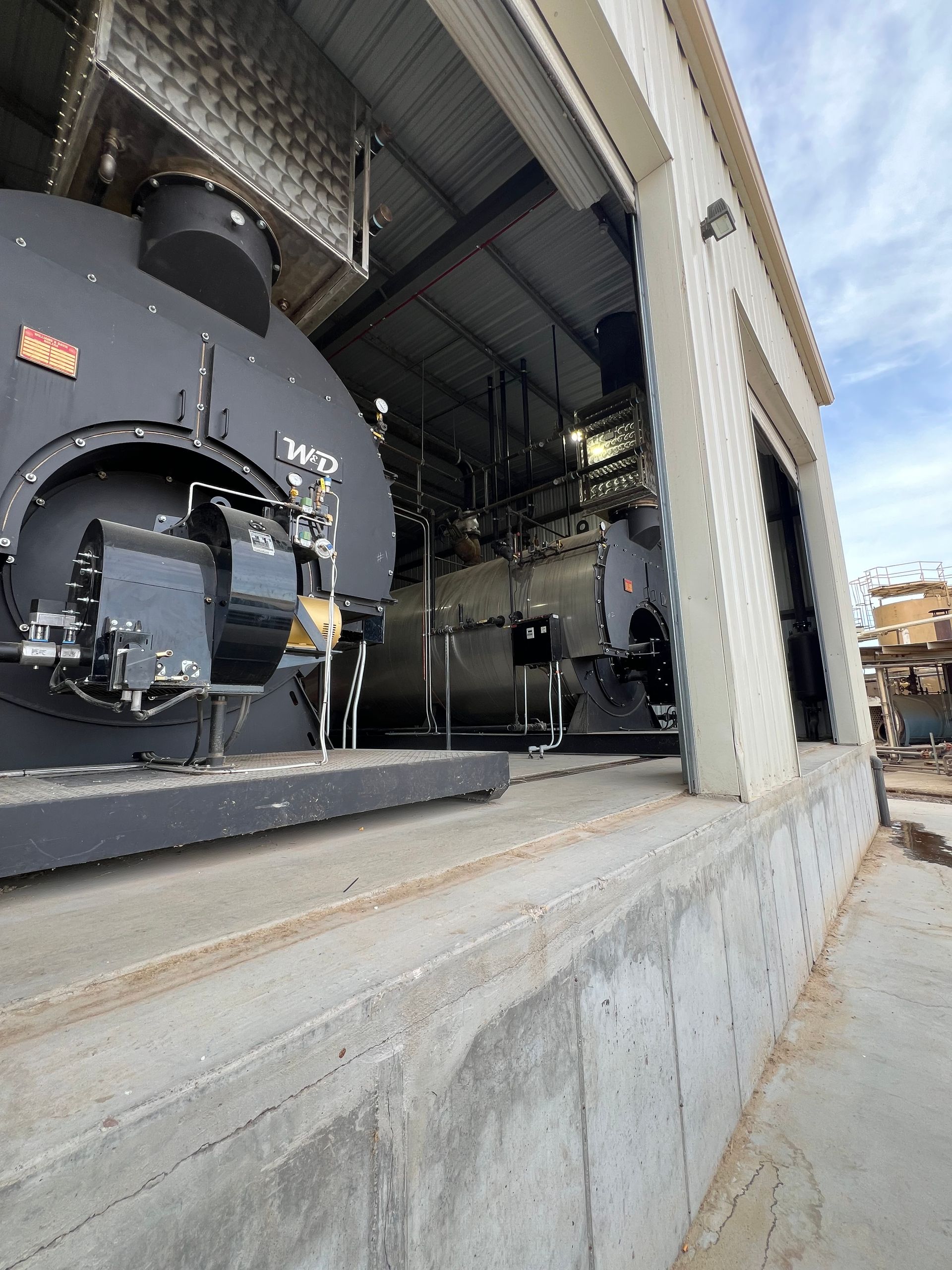 Two large, dark industrial boilers inside a building with open doorway. Concrete foundation, daylight.