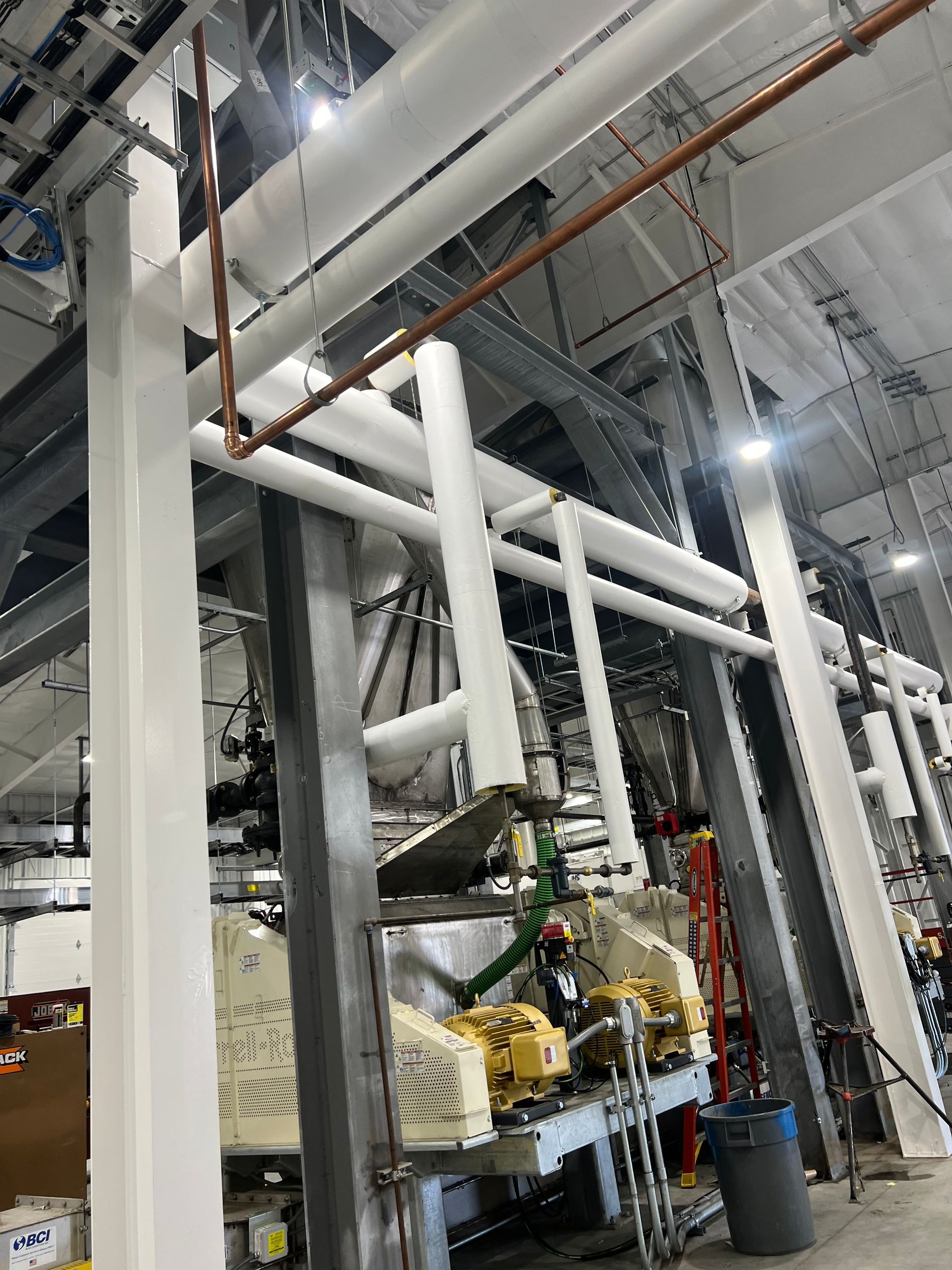 Industrial interior with white pipes, structural beams, and machinery, mostly gray and white.
