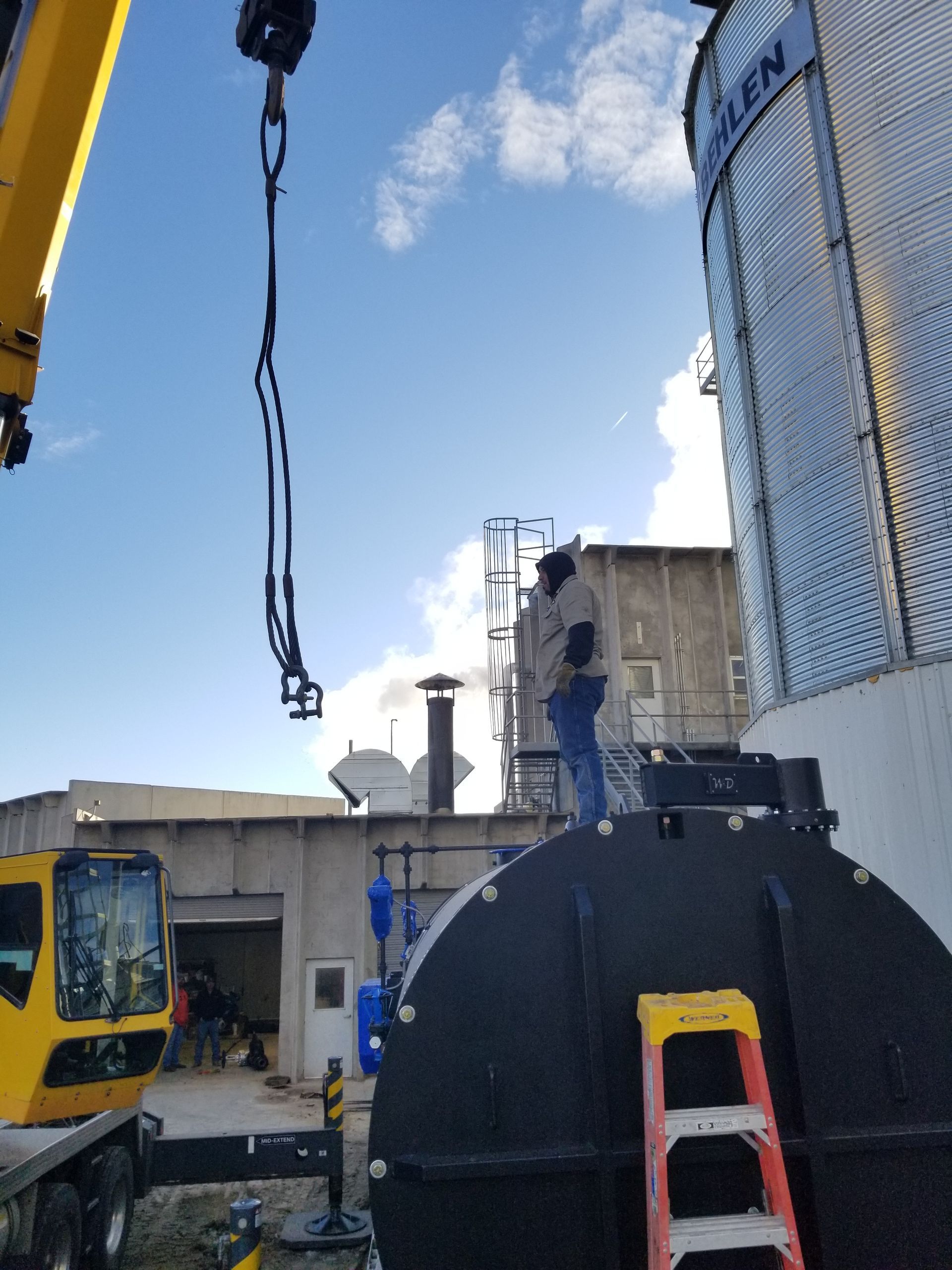 Man atop a black tank near a building, being assisted by a crane. Blue sky, ladder present.