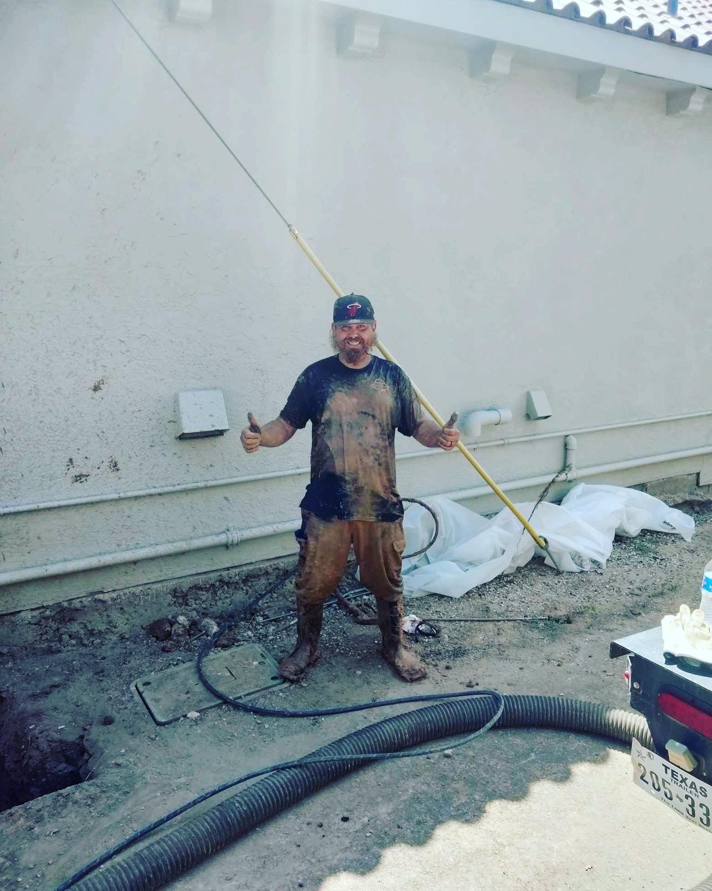 Man covered in mud gives thumbs-up, holding a long pole next to a building.