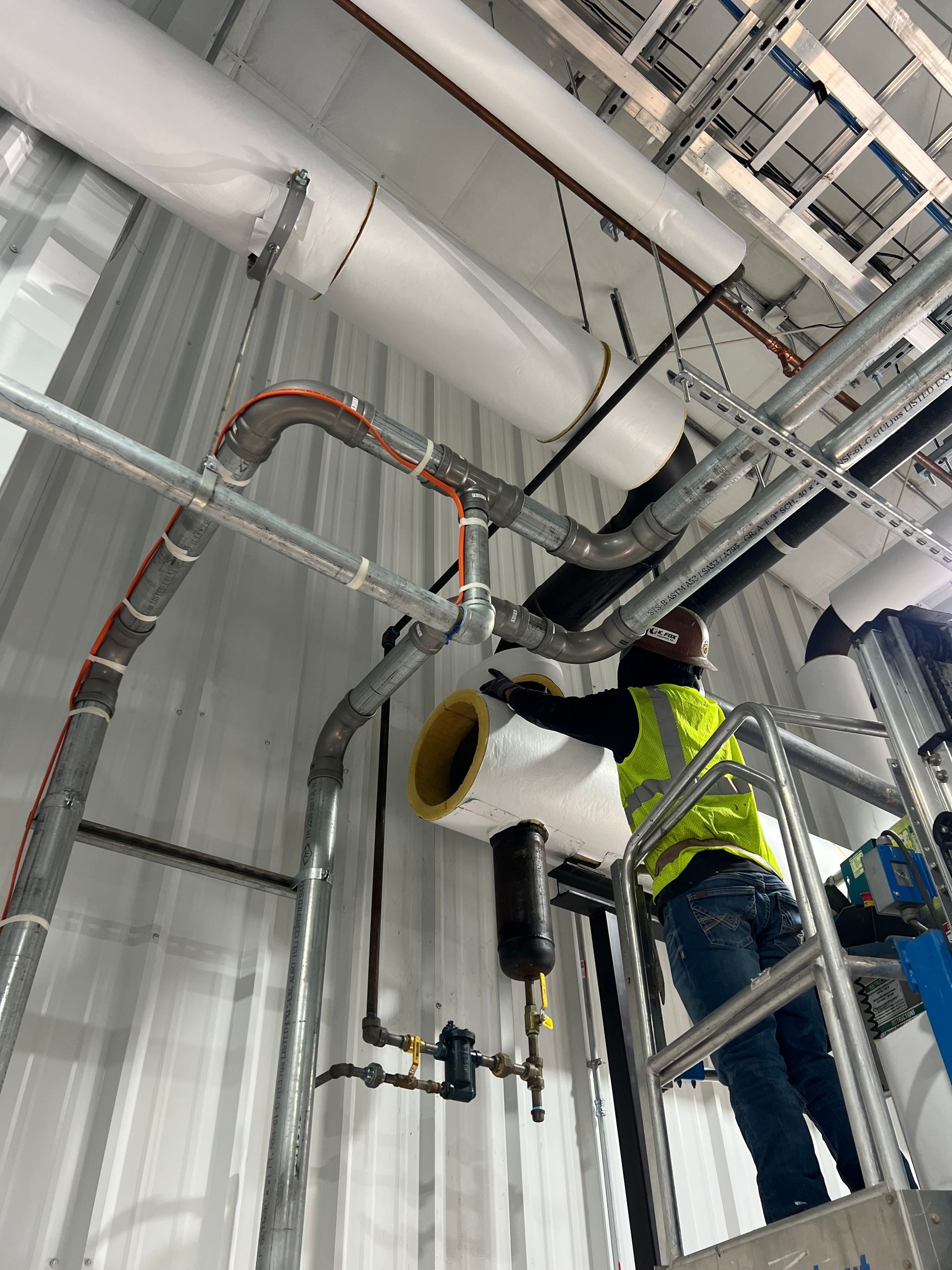 Construction worker on a lift, installing pipes on a ceiling. He wears a yellow vest.