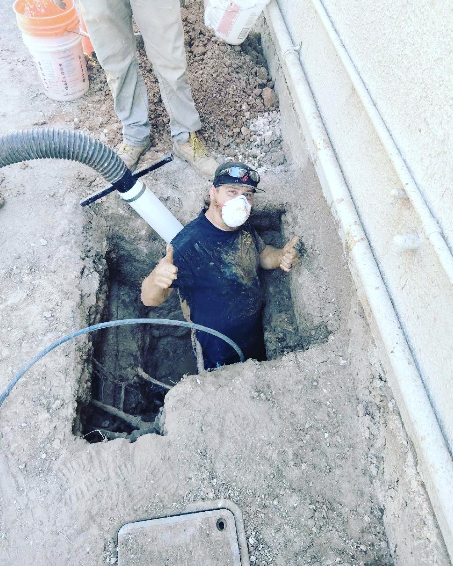 Man in trench giving thumbs up, working on a project near a wall. He wears a mask, dirty clothes.