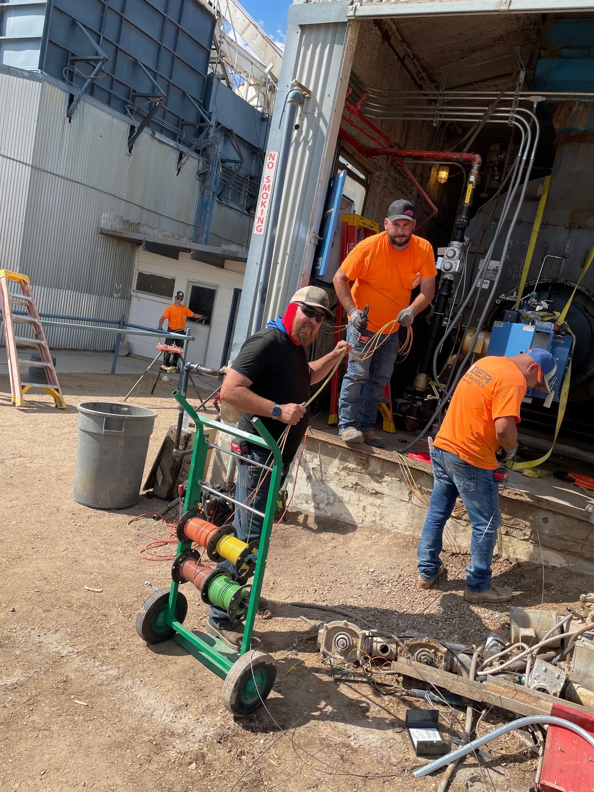 Construction workers in orange shirts work near a building. One man pushes a spool cart.
