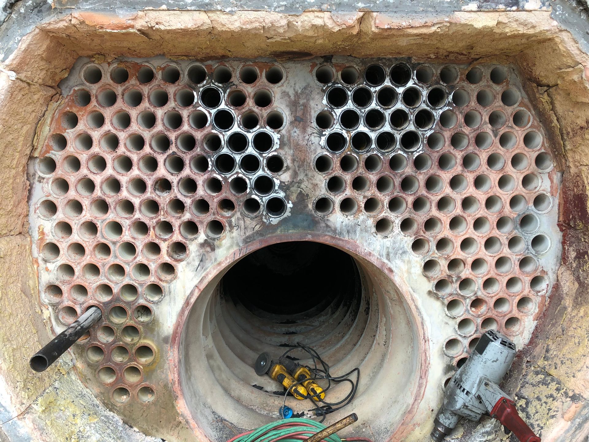 Inside of a boiler with numerous holes. The boiler is round with dark interior and tools resting near the opening.