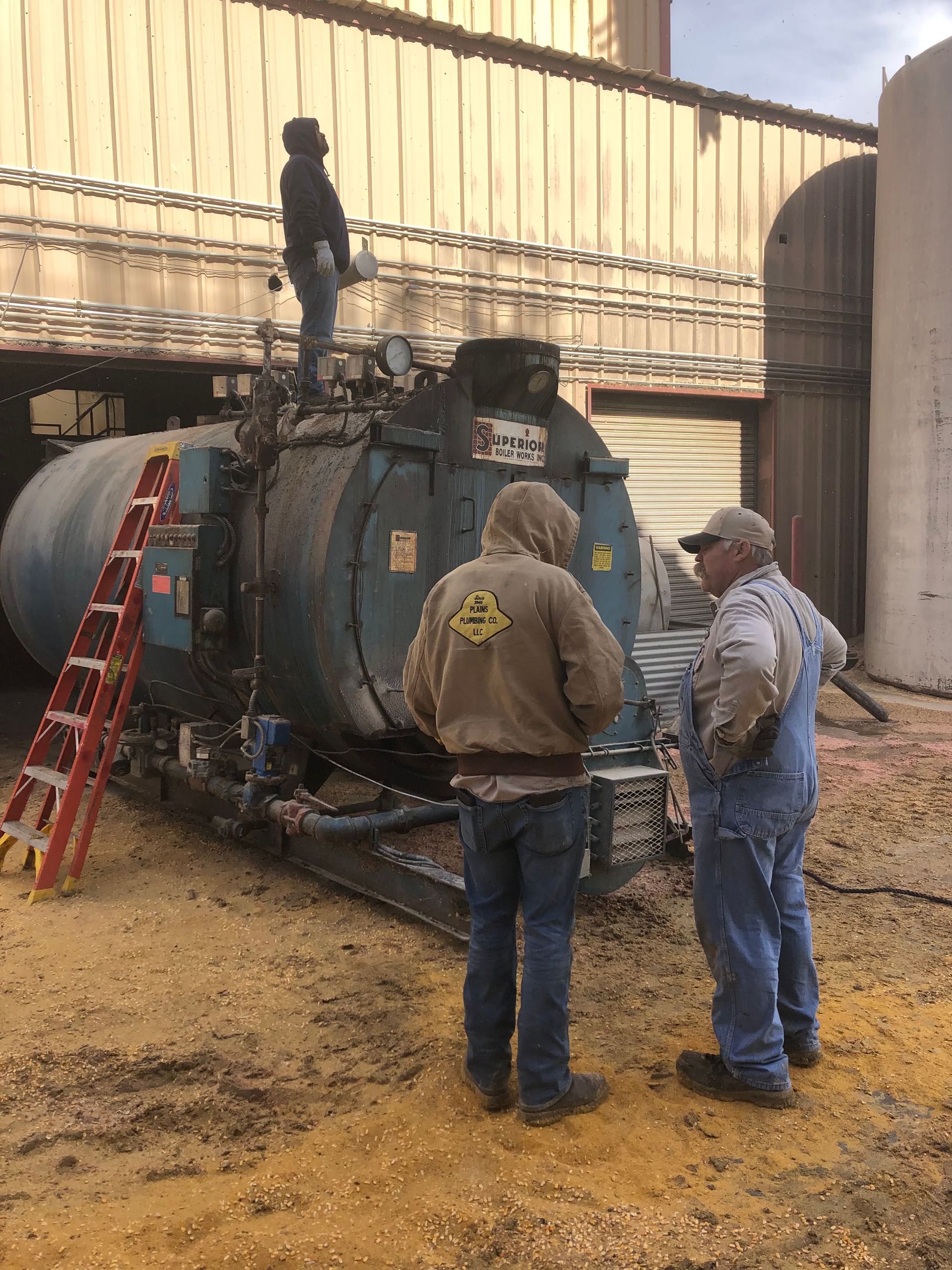 Three men near a blue tank, one atop it. Outdoors, industrial setting.