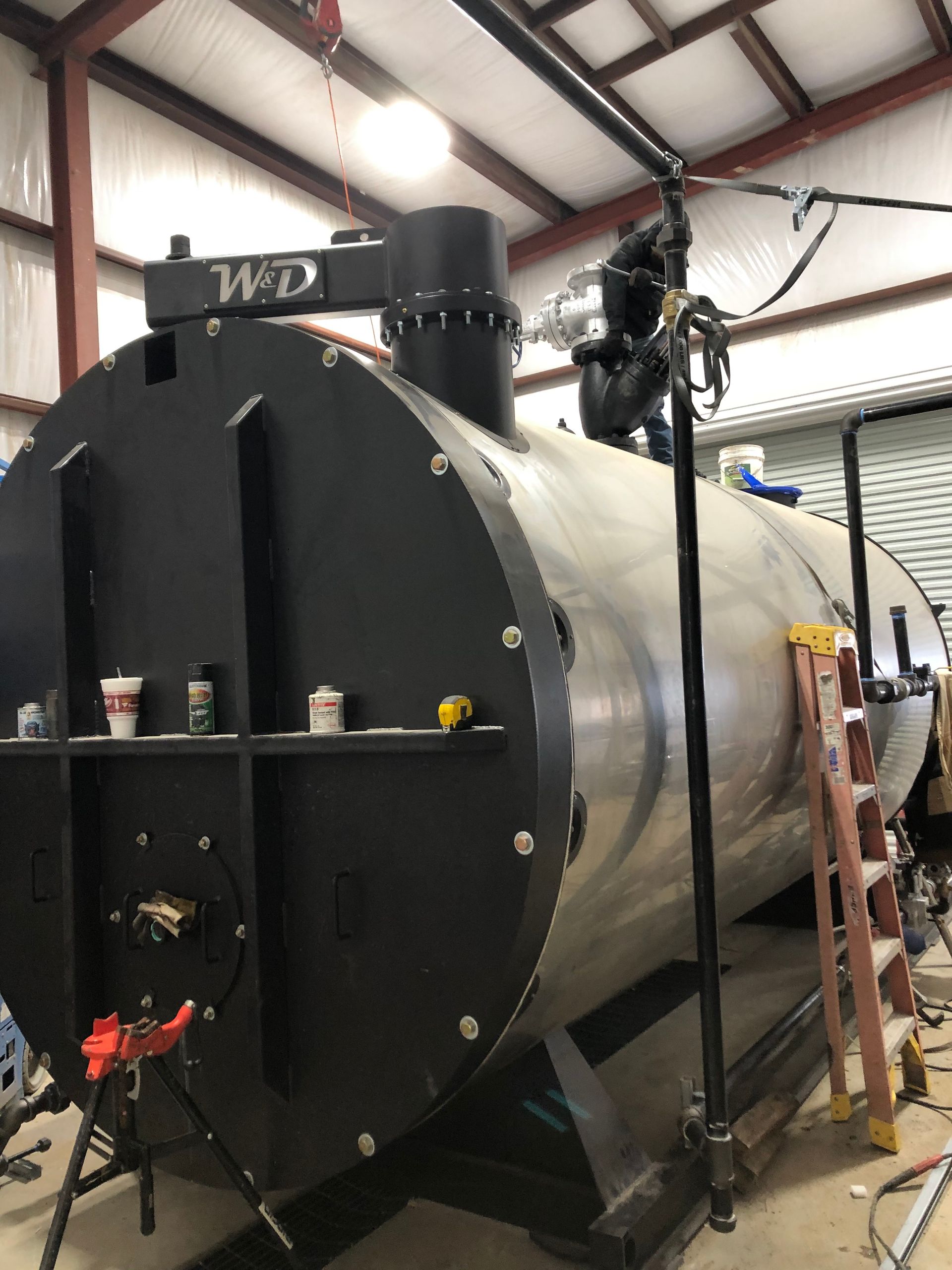 Large cylindrical tank with black ends and a silver body, indoors, with a ladder and a person on top.