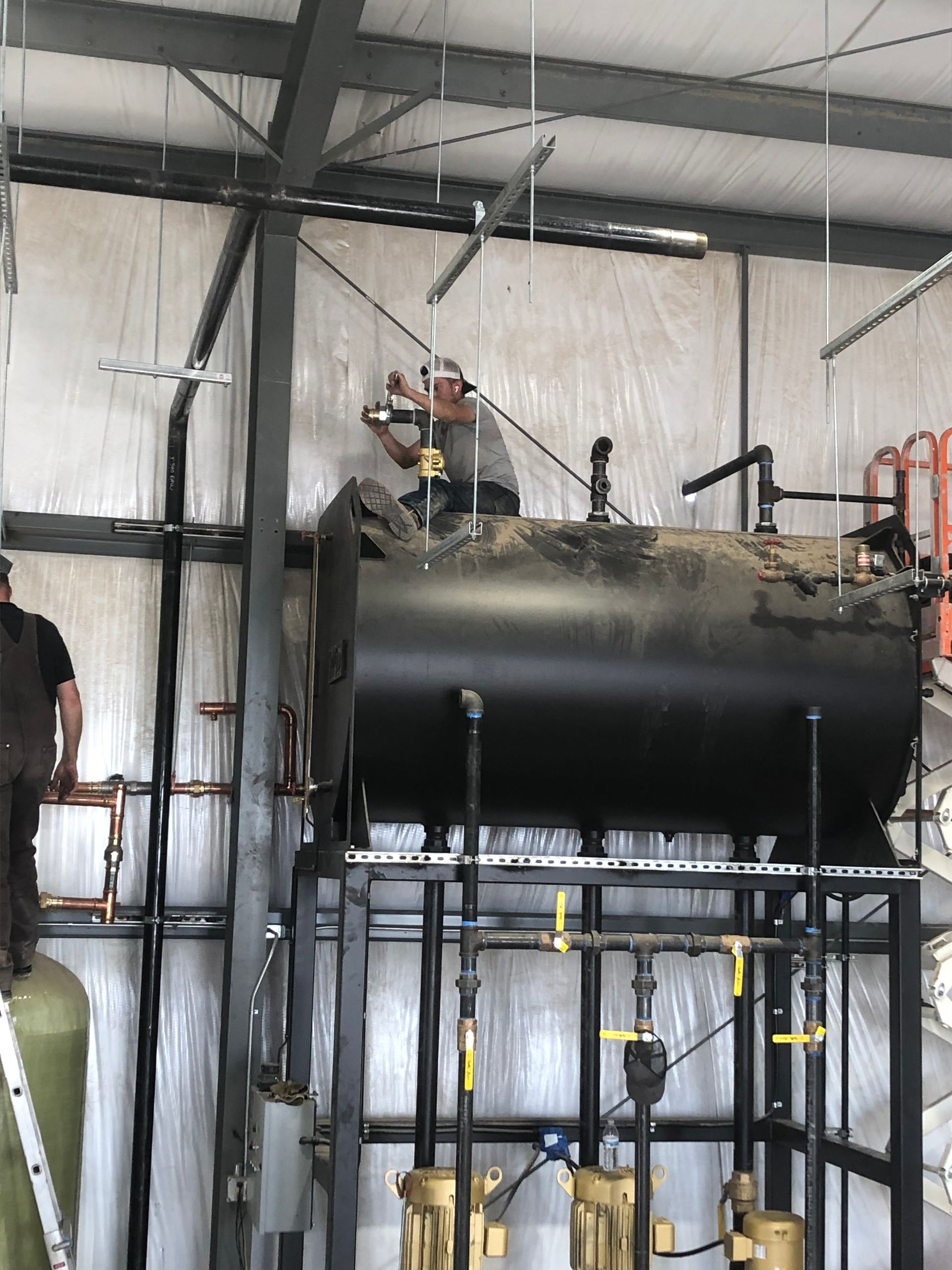 Two men working on a large black tank inside a building, likely industrial; one on top, one on the left side.