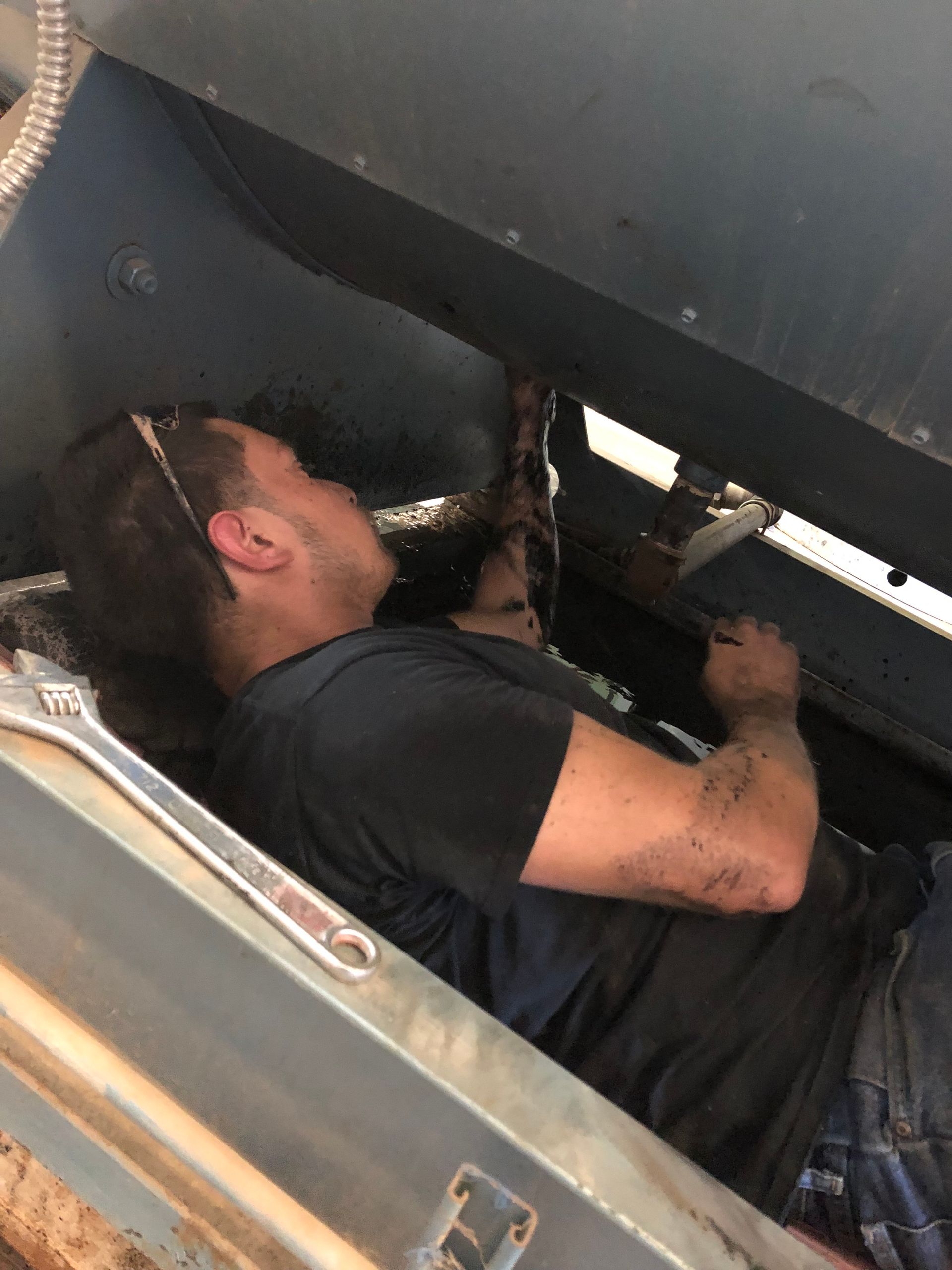 Man lying under machinery, reaching up to work with a wrench; dirty clothes and hands.