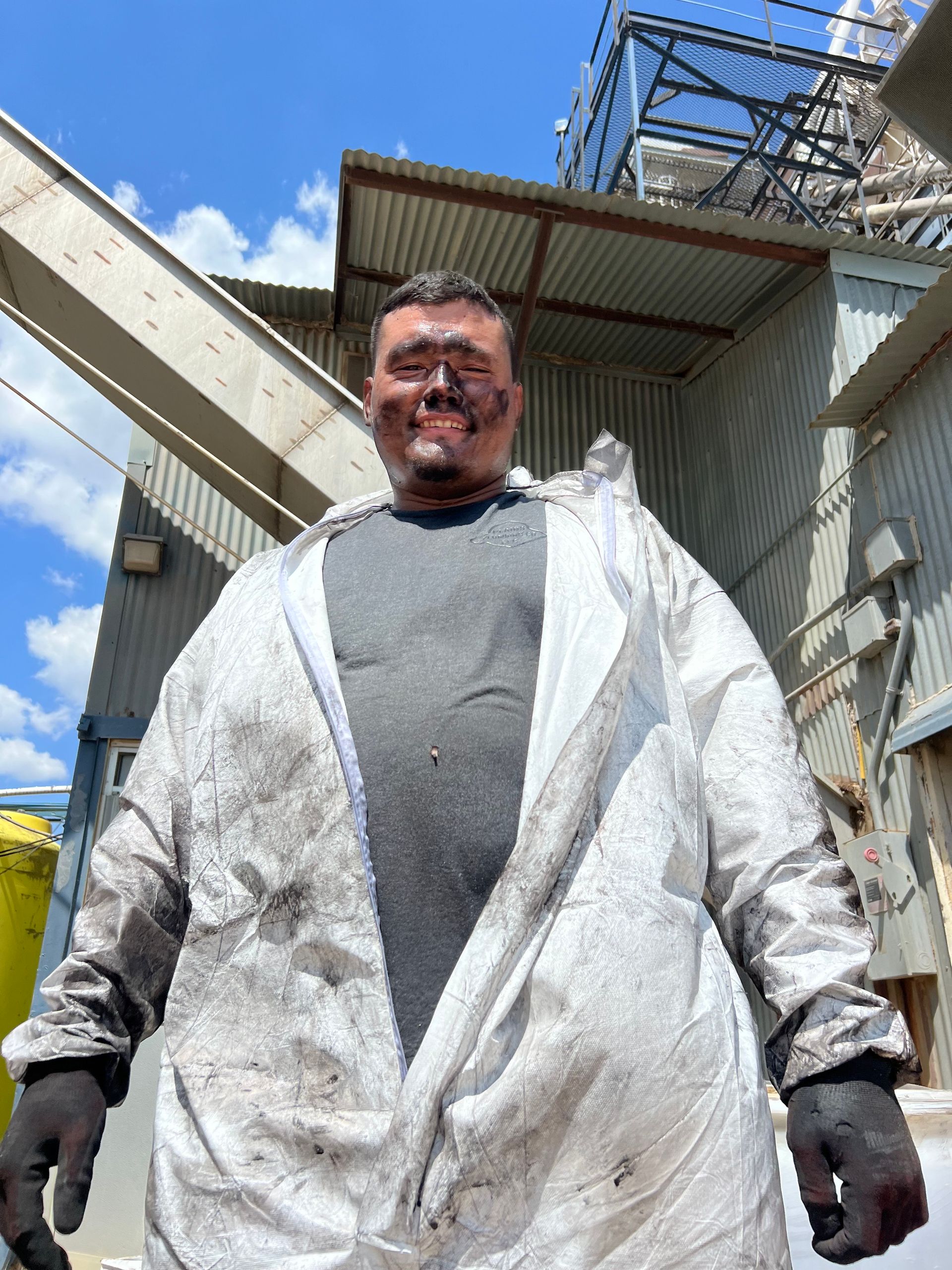 Man in soiled white jumpsuit with soot on face, stands near industrial building.