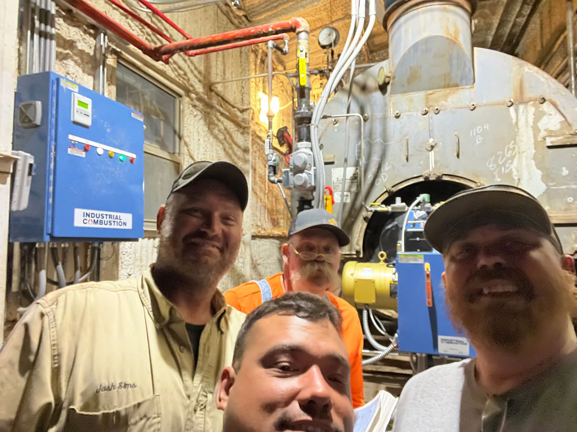 Four men pose in front of industrial equipment in a dimly lit room. They appear to be smiling.