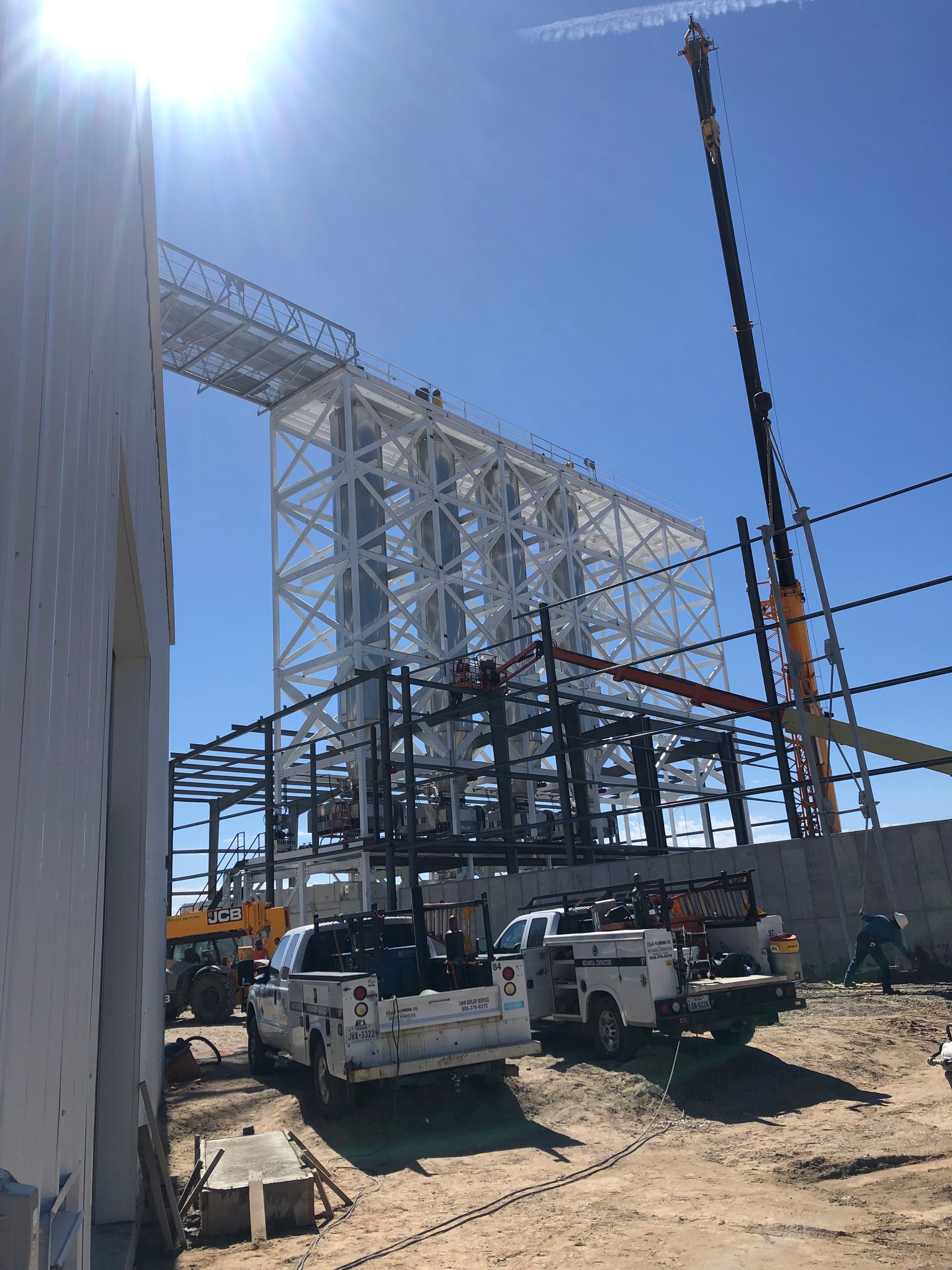 Construction site with white steel framework under blue sky, trucks, crane, and workers.