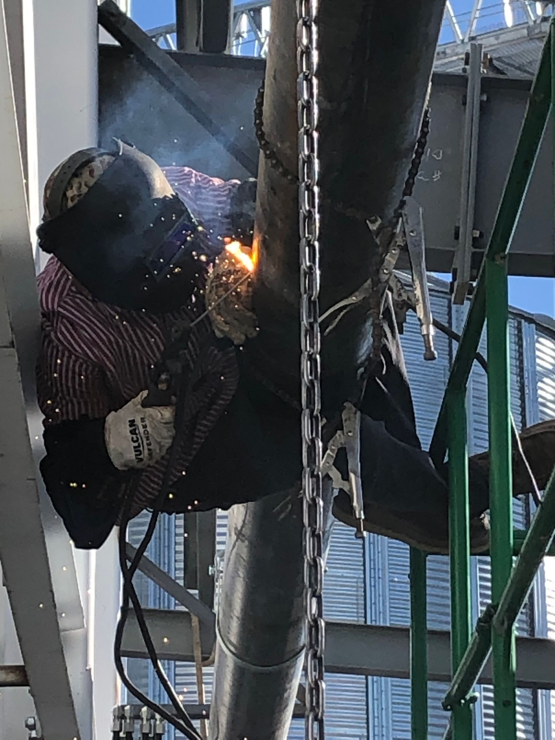 Welder in protective gear welds a pipe high above ground. Sparks fly.