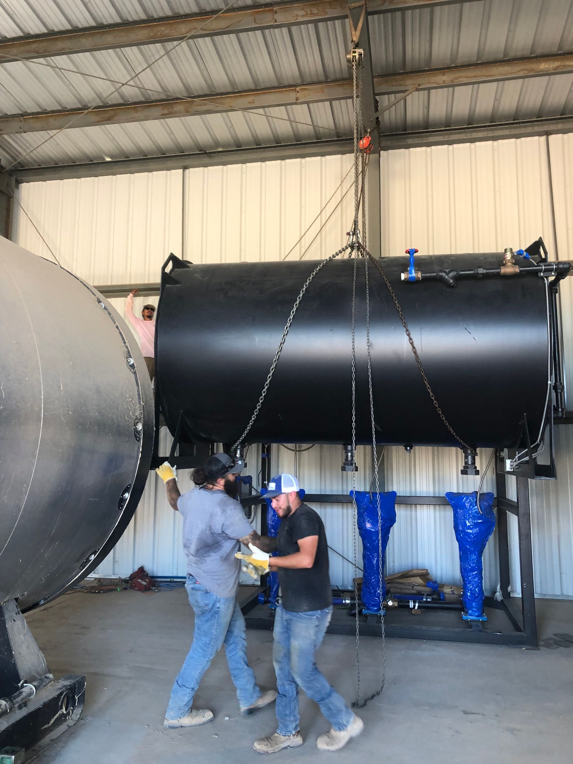 Two men installing a black tank, suspended by a crane, inside a metal building.
