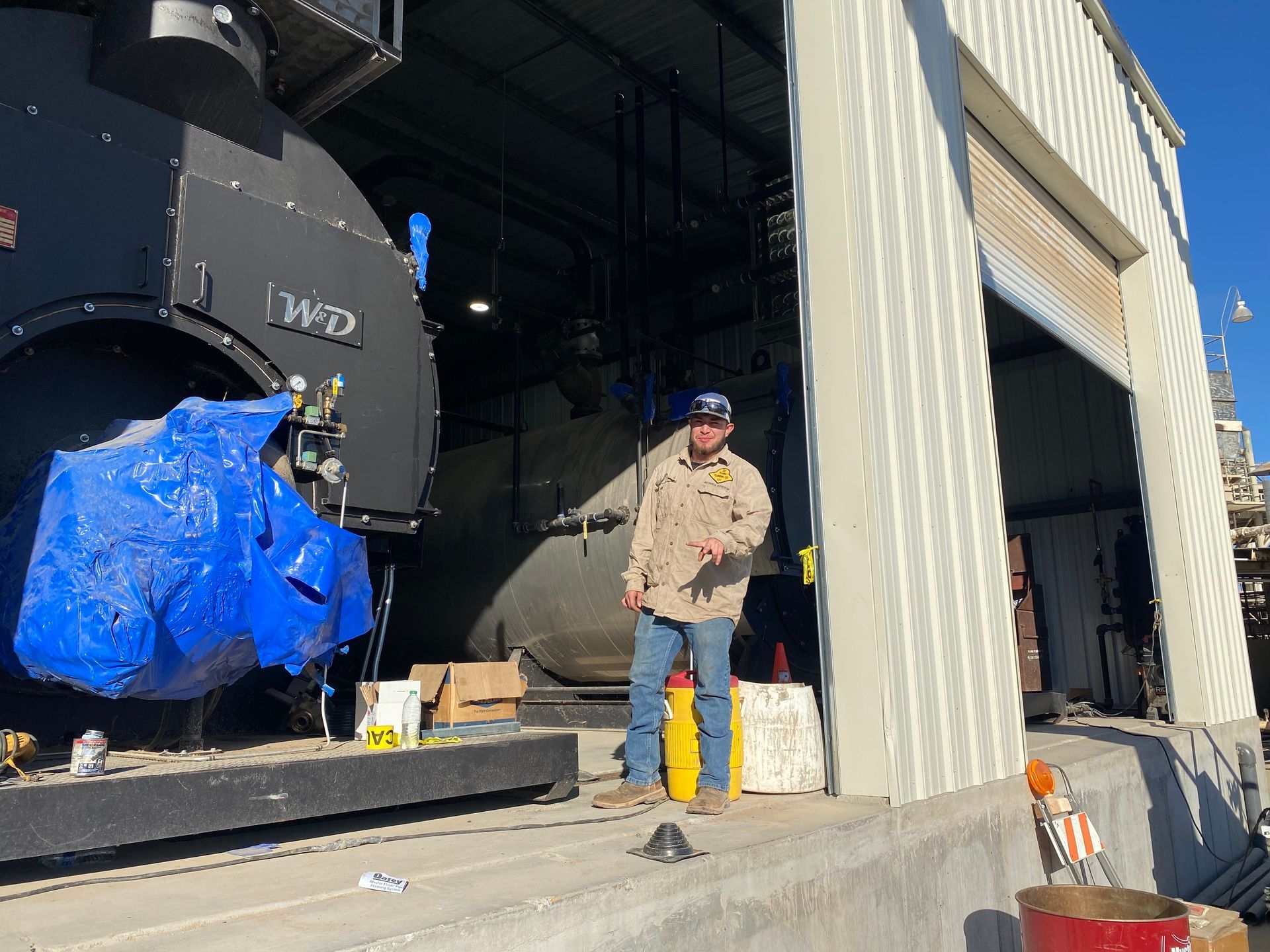 Man stands near industrial equipment in a building. He wears jeans and a work shirt, holding a pen.
