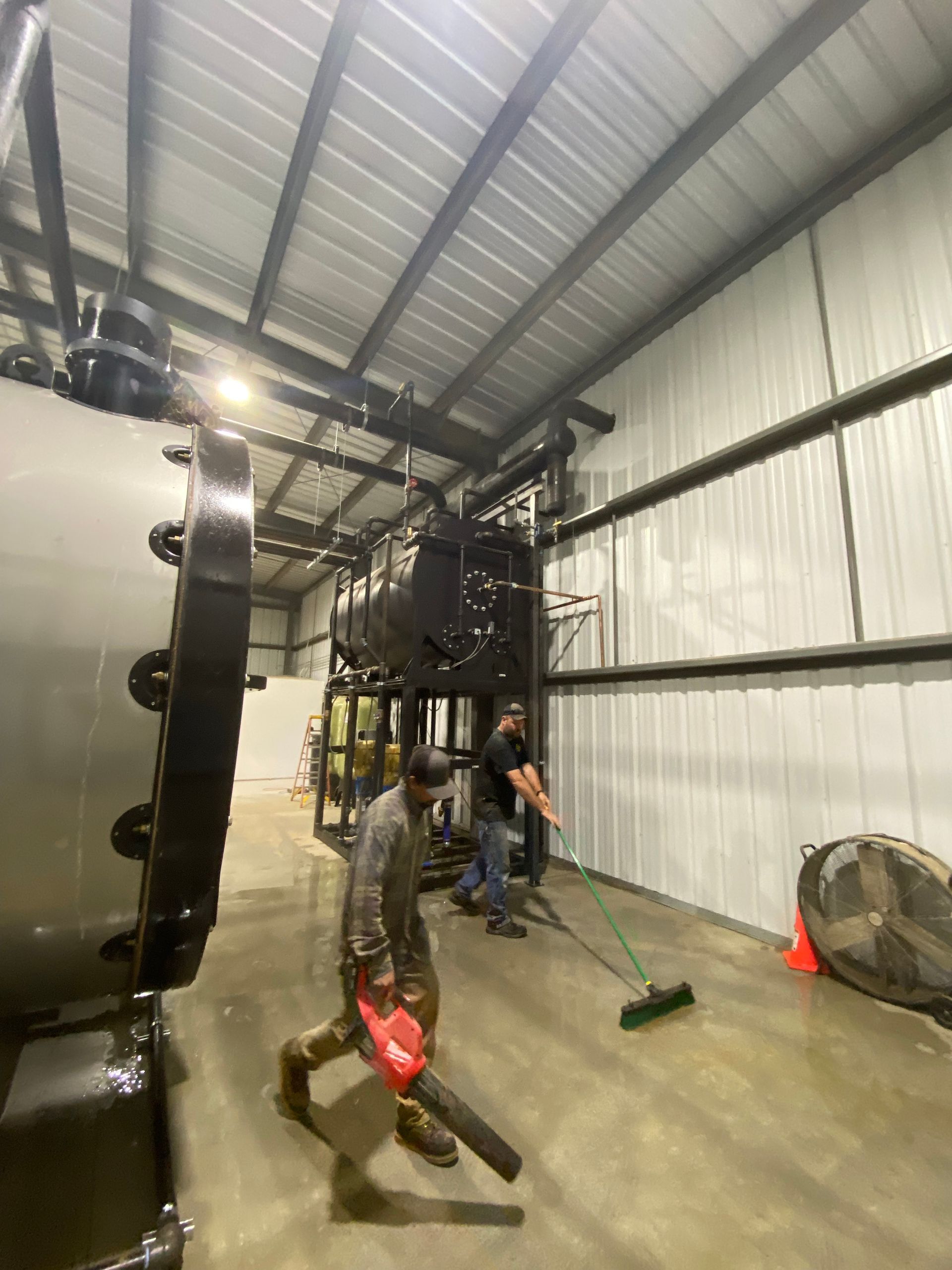 Men cleaning a warehouse with a leaf blower and broom. Steel tanks and walls visible.