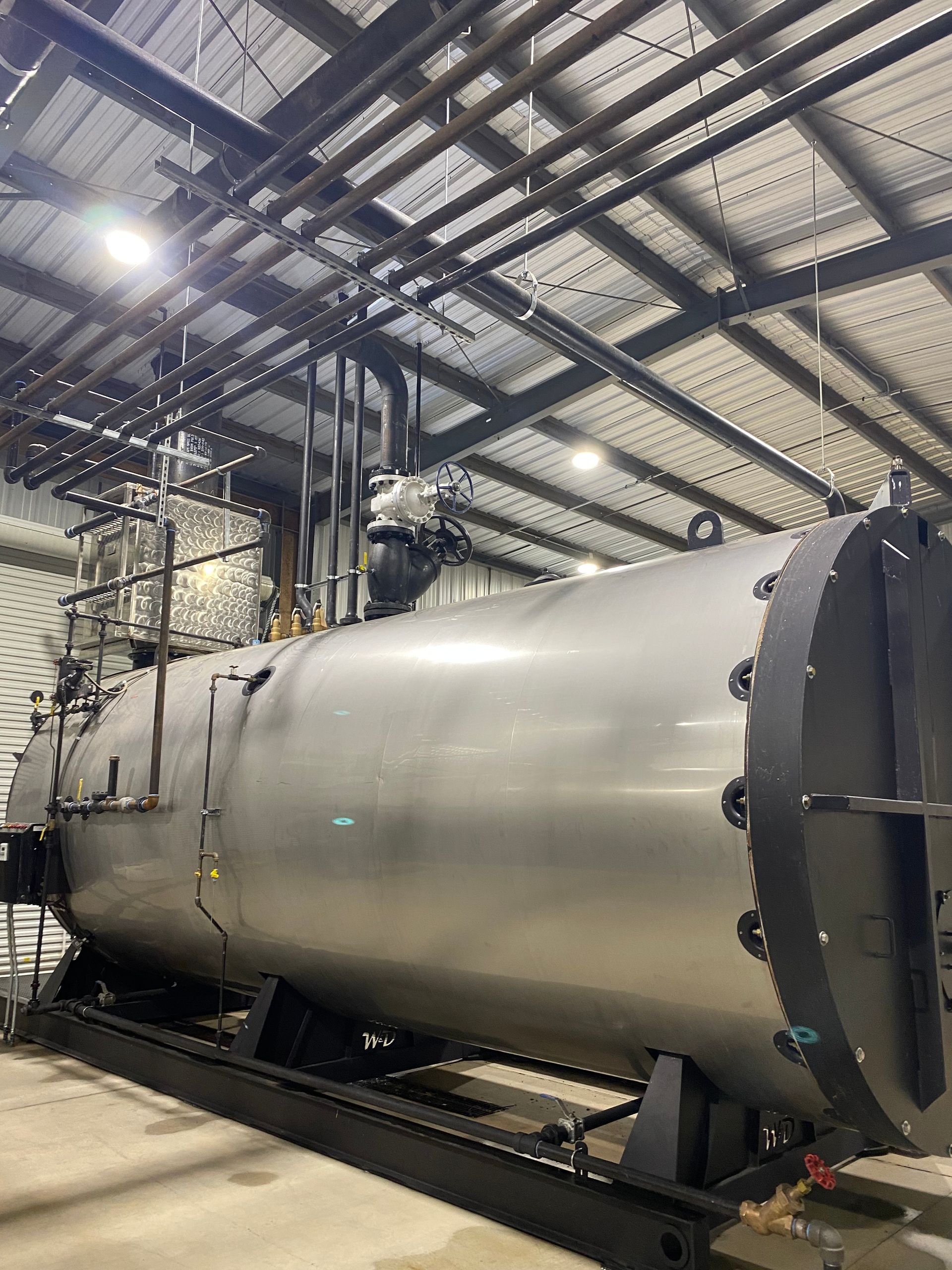 A large, silver industrial boiler in a metal building. Pipes run along the ceiling and connect to the boiler.