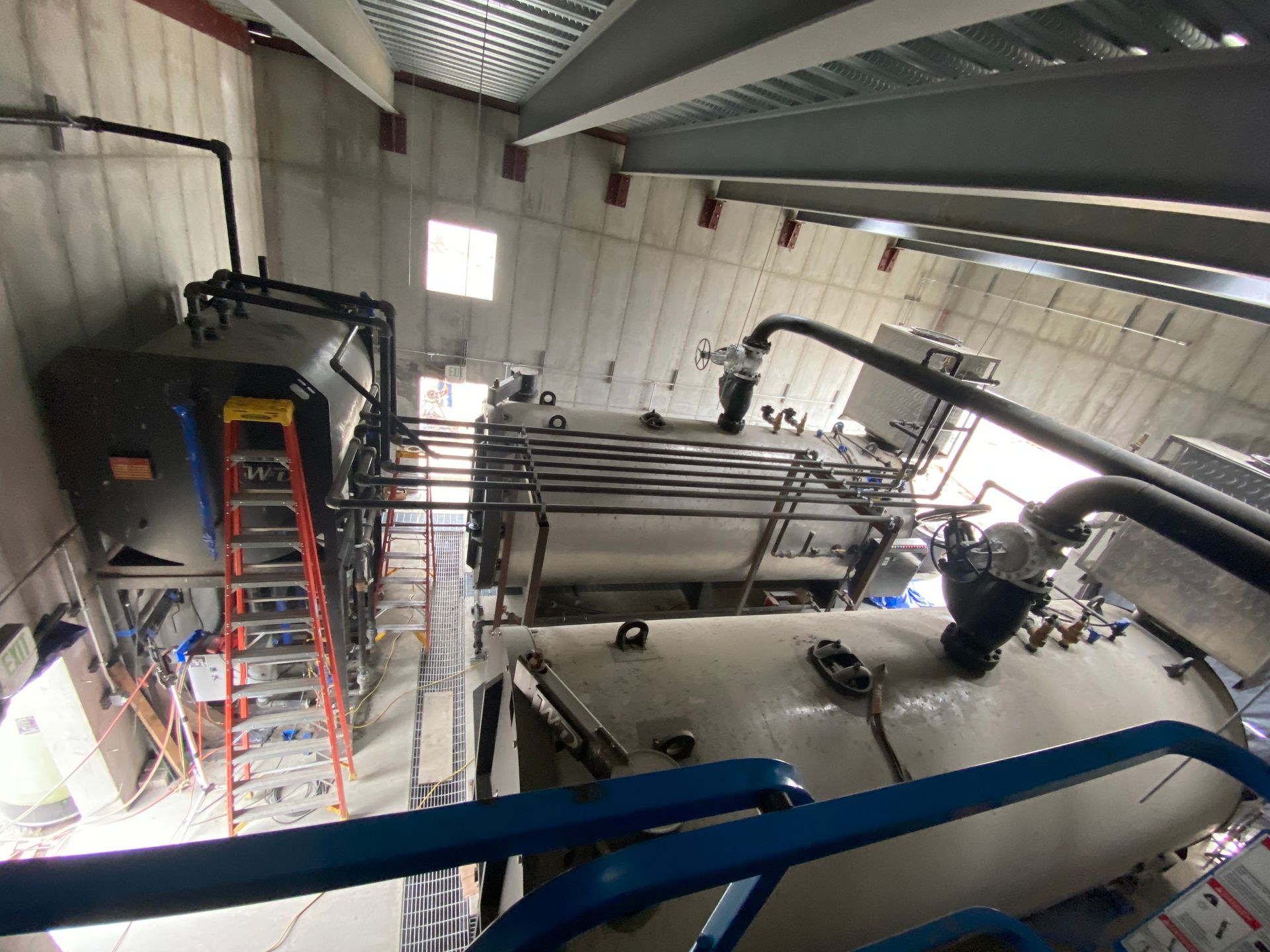 Industrial equipment in a building, featuring tanks, pipes, and a ladder. Overhead view, mostly gray with black pipes.