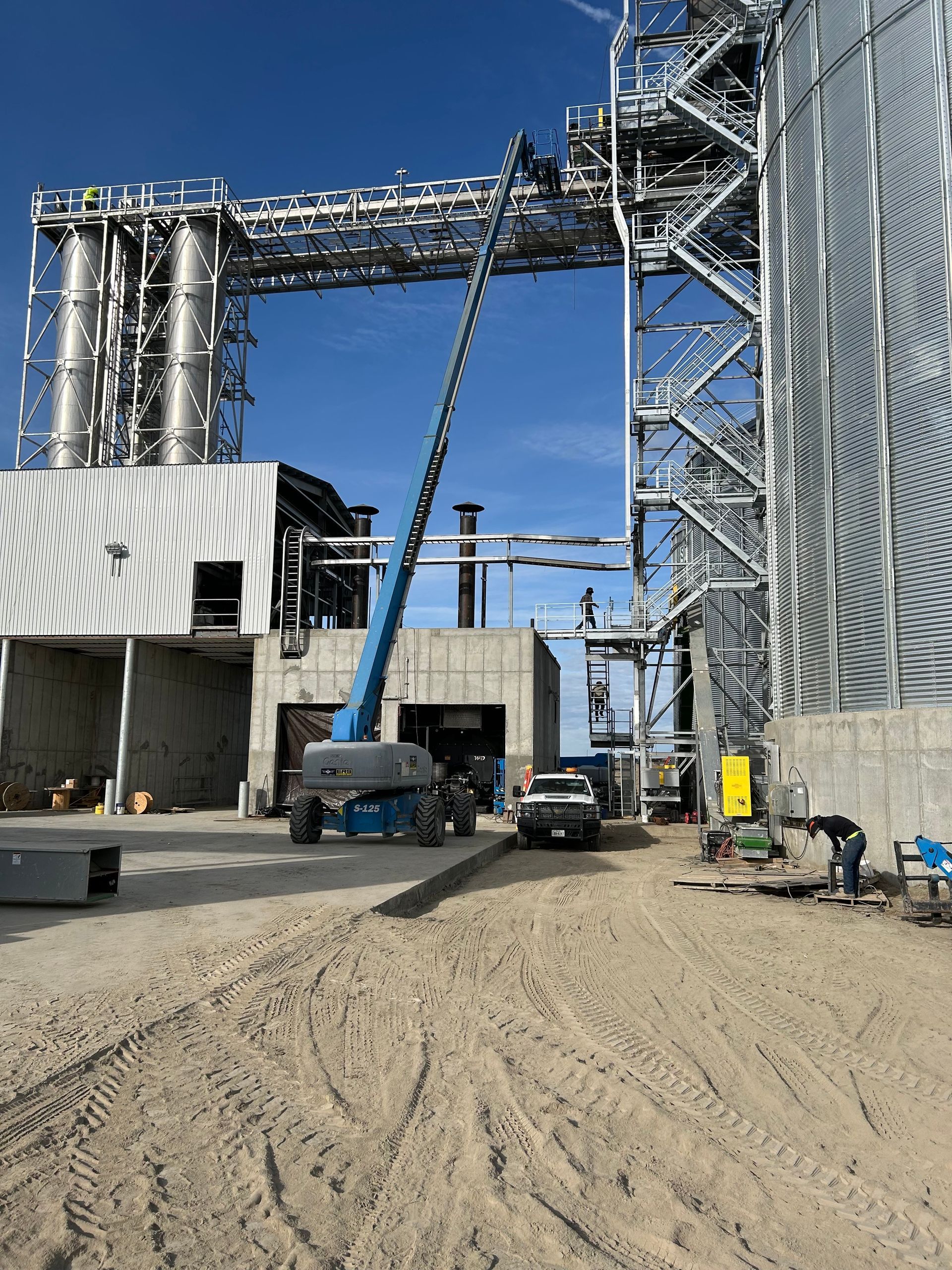 Construction site with a blue lift. Large industrial building with metal silos and pipes under a blue sky.