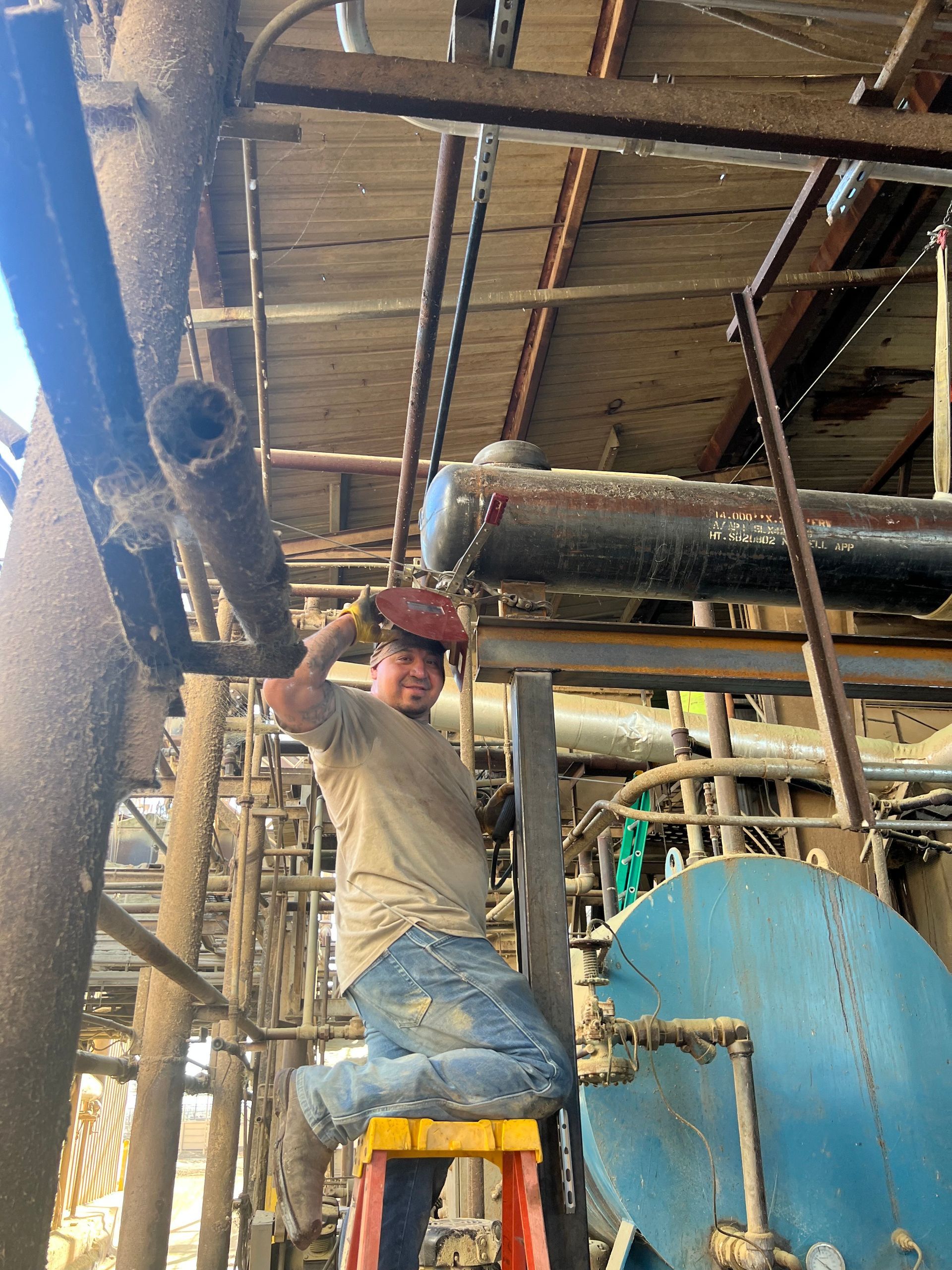 A worker wearing a welding helmet welds pipes on an industrial structure while kneeling on a ladder.