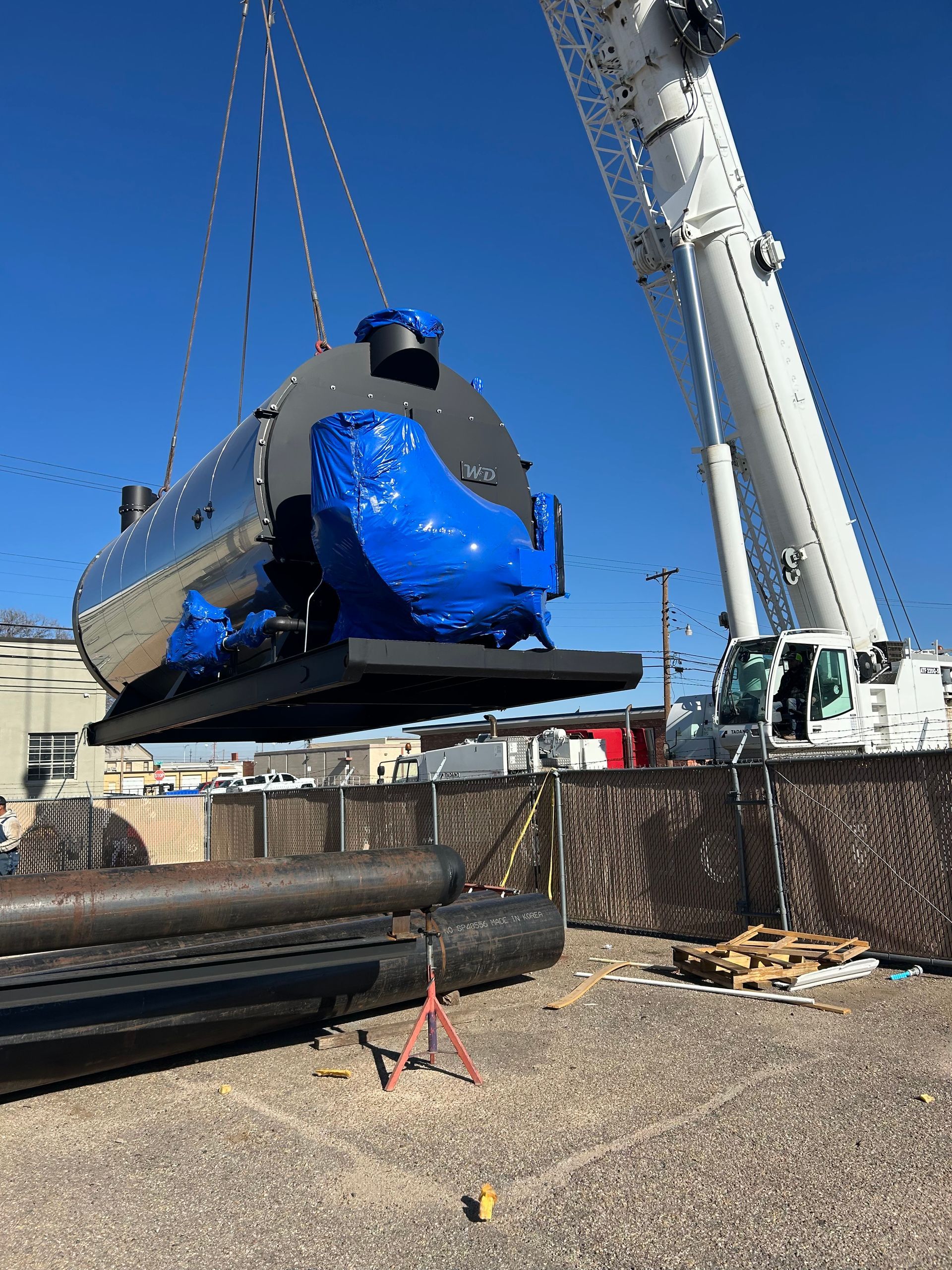 A large, shiny industrial boiler being lifted by a white crane; blue tarp covers the ends, outdoors on a construction site.