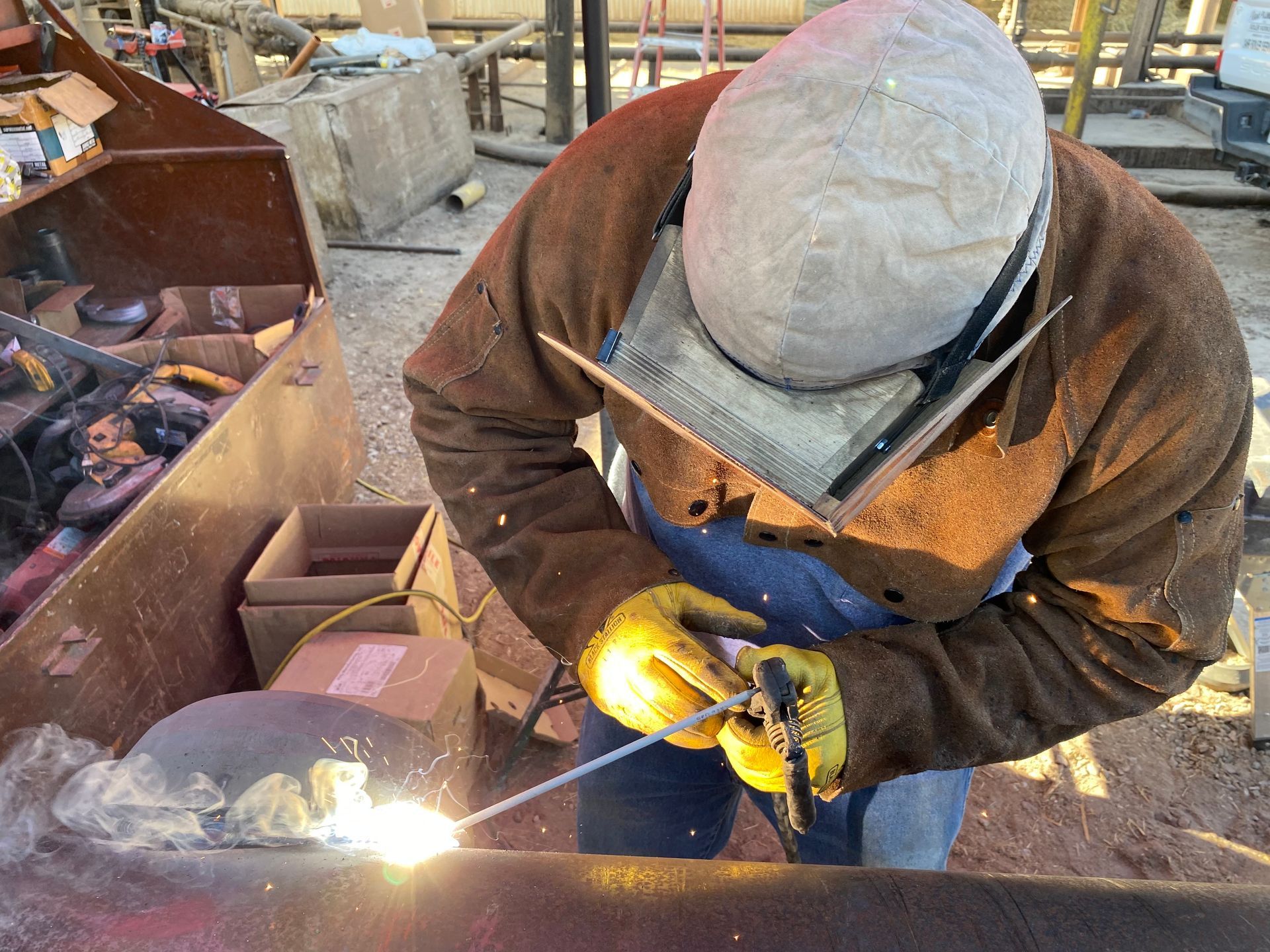 Welder wearing protective gear welding a metal pipe outdoors.