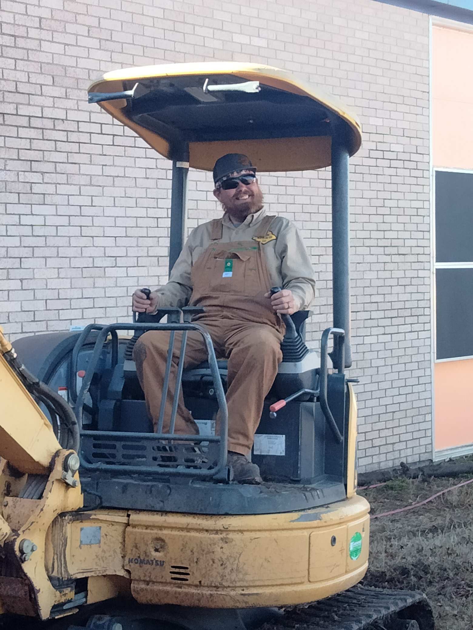 Man in brown overalls operating a yellow excavator in front of a brick building.