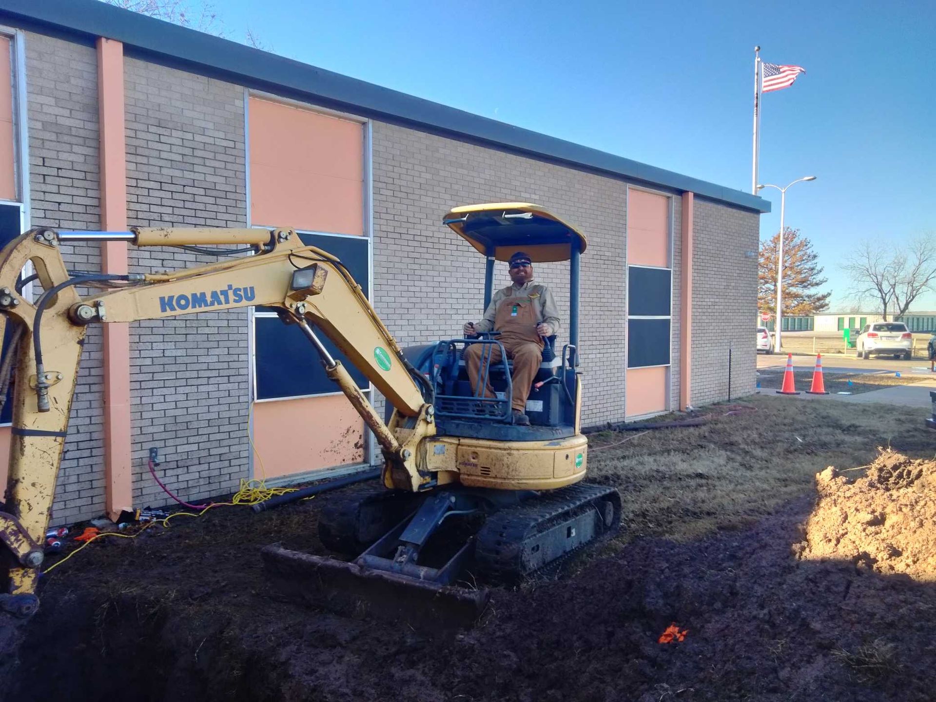 Person operating a yellow Komatsu excavator digging near a school building with an American flag.